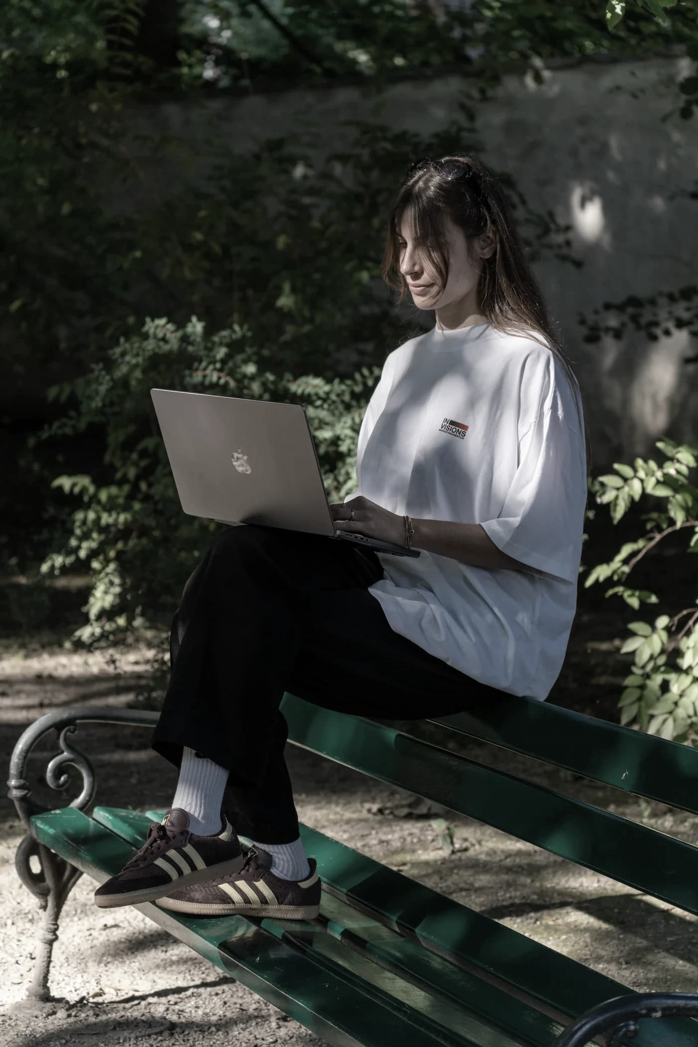 Person mit Invisions-Shirt sitzt mit dem Laptop auf einer Parkbank im Grünen.