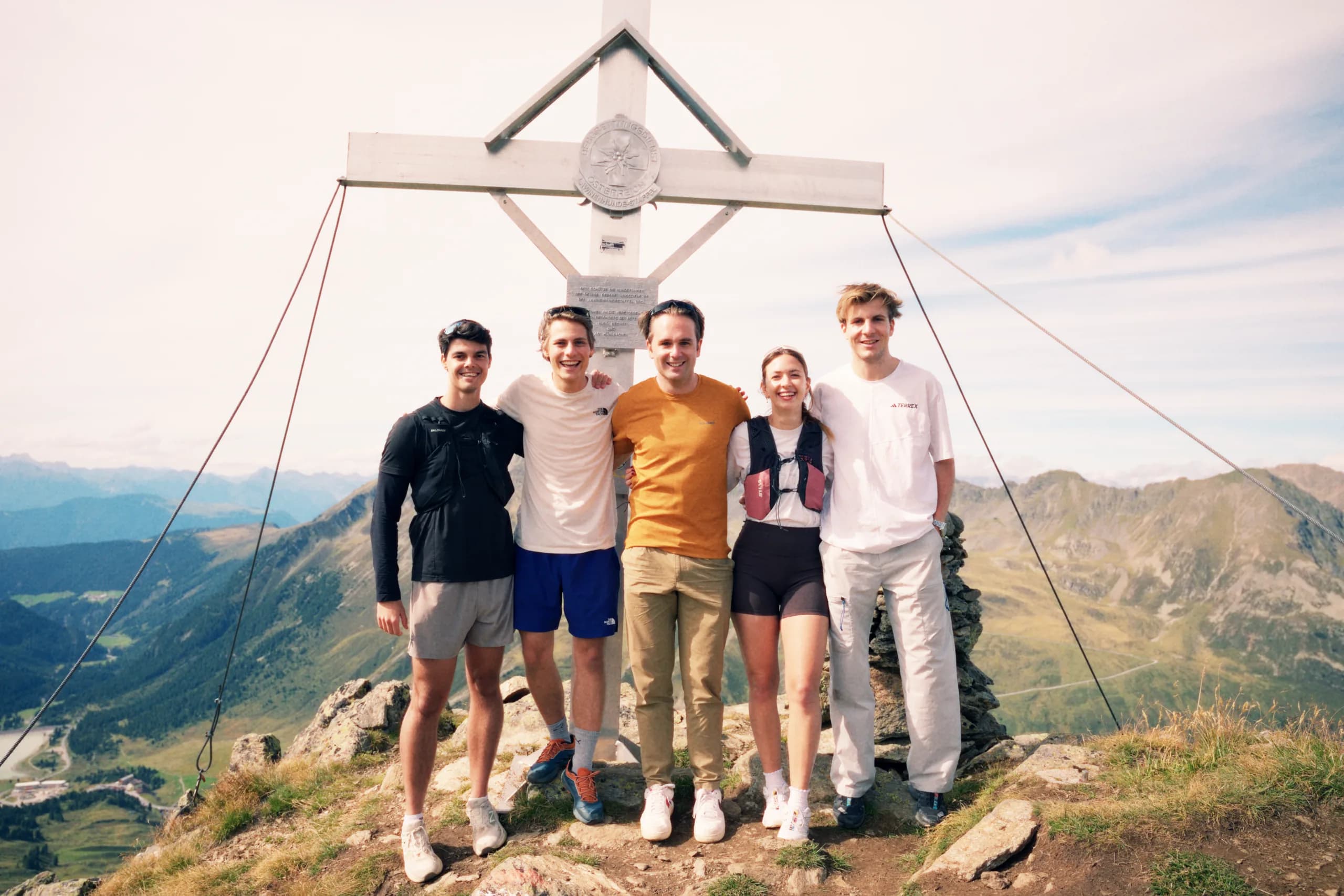Fünf aus dem Team stehen im Sommer nebeneinander vor einem Gipfelkreuz, dahinter das Tal und die Berge gegenüber.