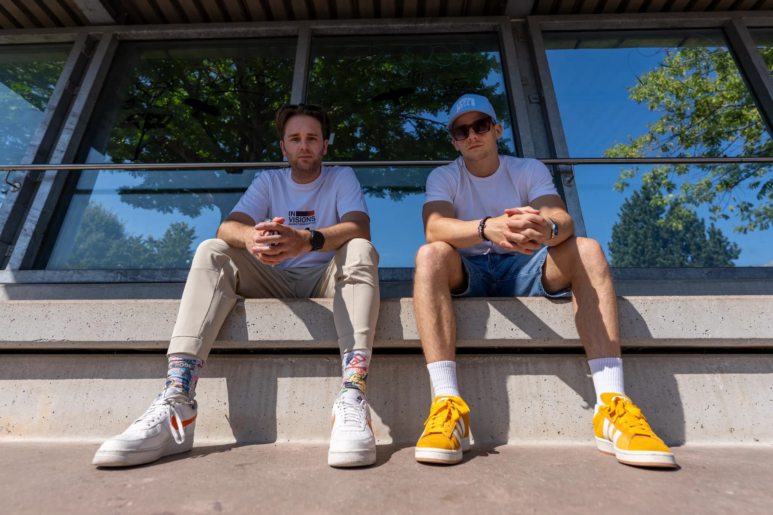 Zwei aus dem Team sitzen im Sommer auf einer Betonstufe vor der Glasfassade des Büros.