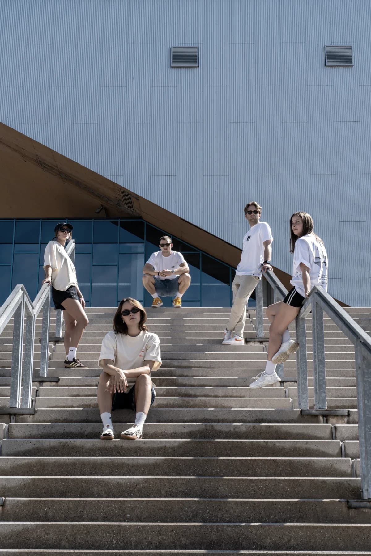 Fünf aus dem Invisions-Team auf einer breiten Außentreppe vor einer hellen Metallfassade, in weißen Shirts und Sonnenbrillen im Mittagslicht.