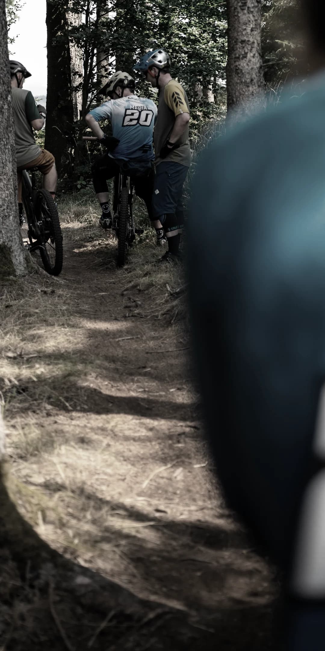 Mountainbiker auf einem Waldtrail, vorn unscharf ein Fahrer im Invisions-Trikot.