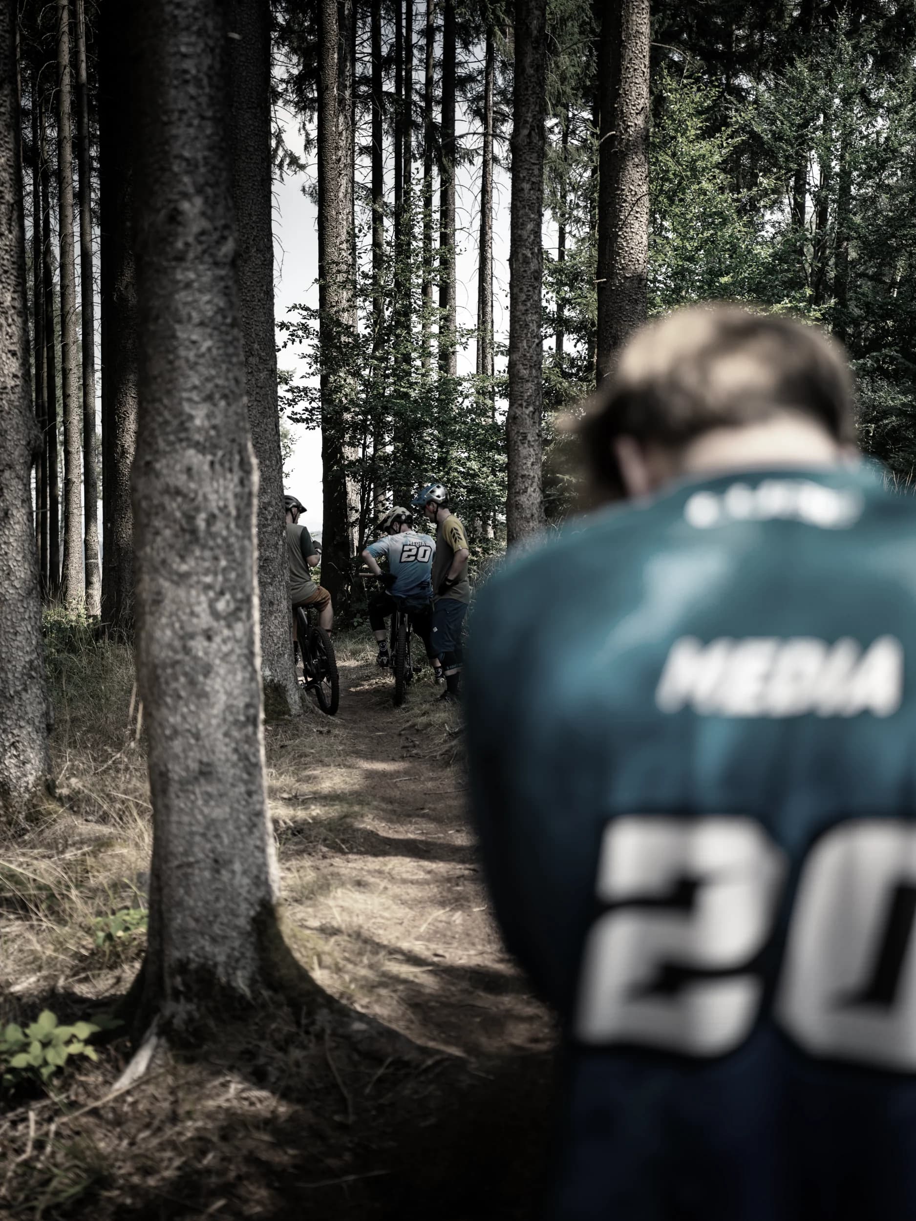 Mountainbiker auf einem Waldtrail, vorn unscharf ein Fahrer im Invisions-Trikot.