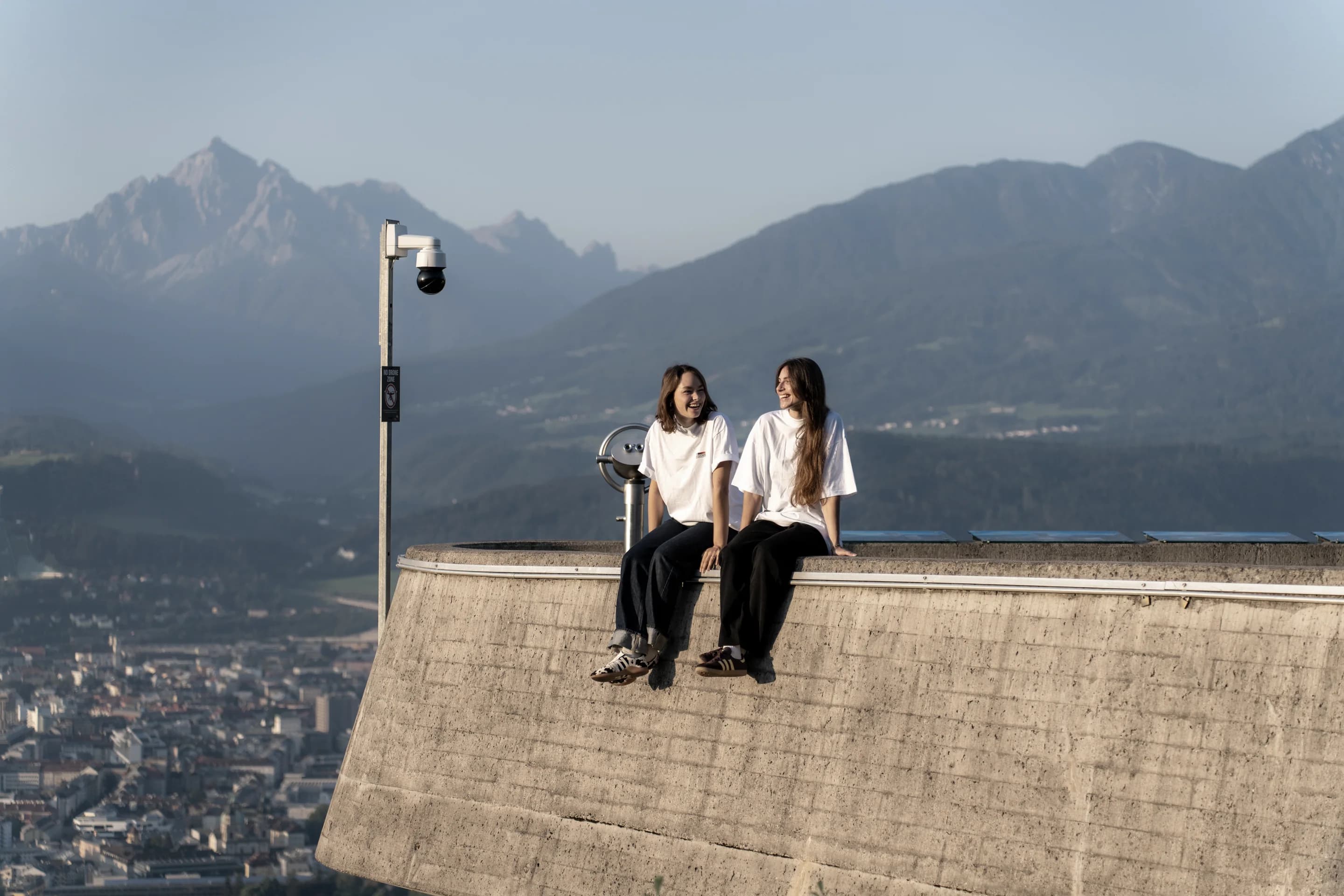 Zwei aus dem Team sitzen in Invisions-Shirts auf der Brüstung am Bergisel, dahinter Innsbruck und die Berge.