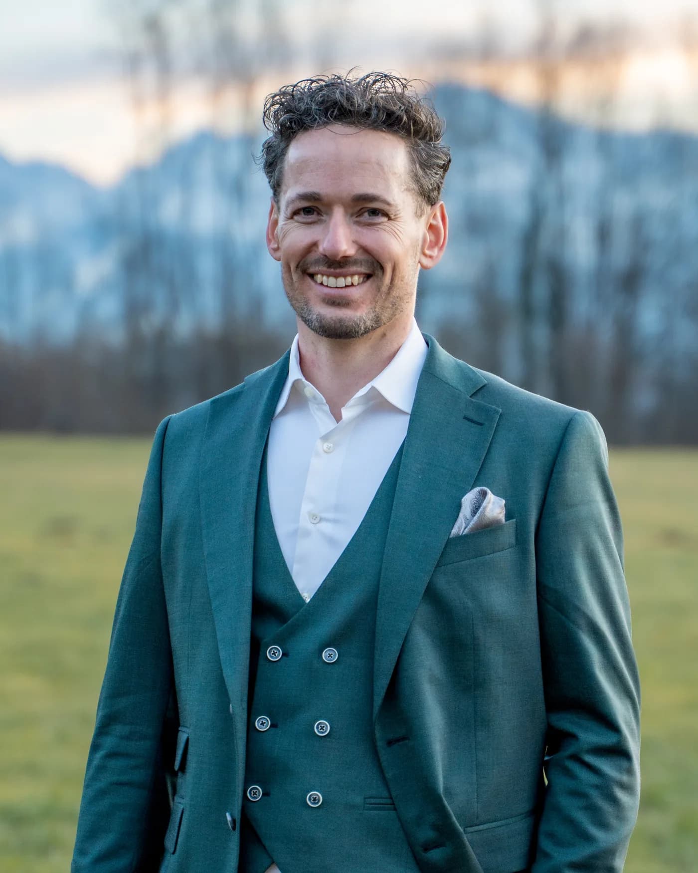 Florian Brucker im grünen Anzug auf der Wiese, lächelnd, dahinter die Berge im Abendlicht.