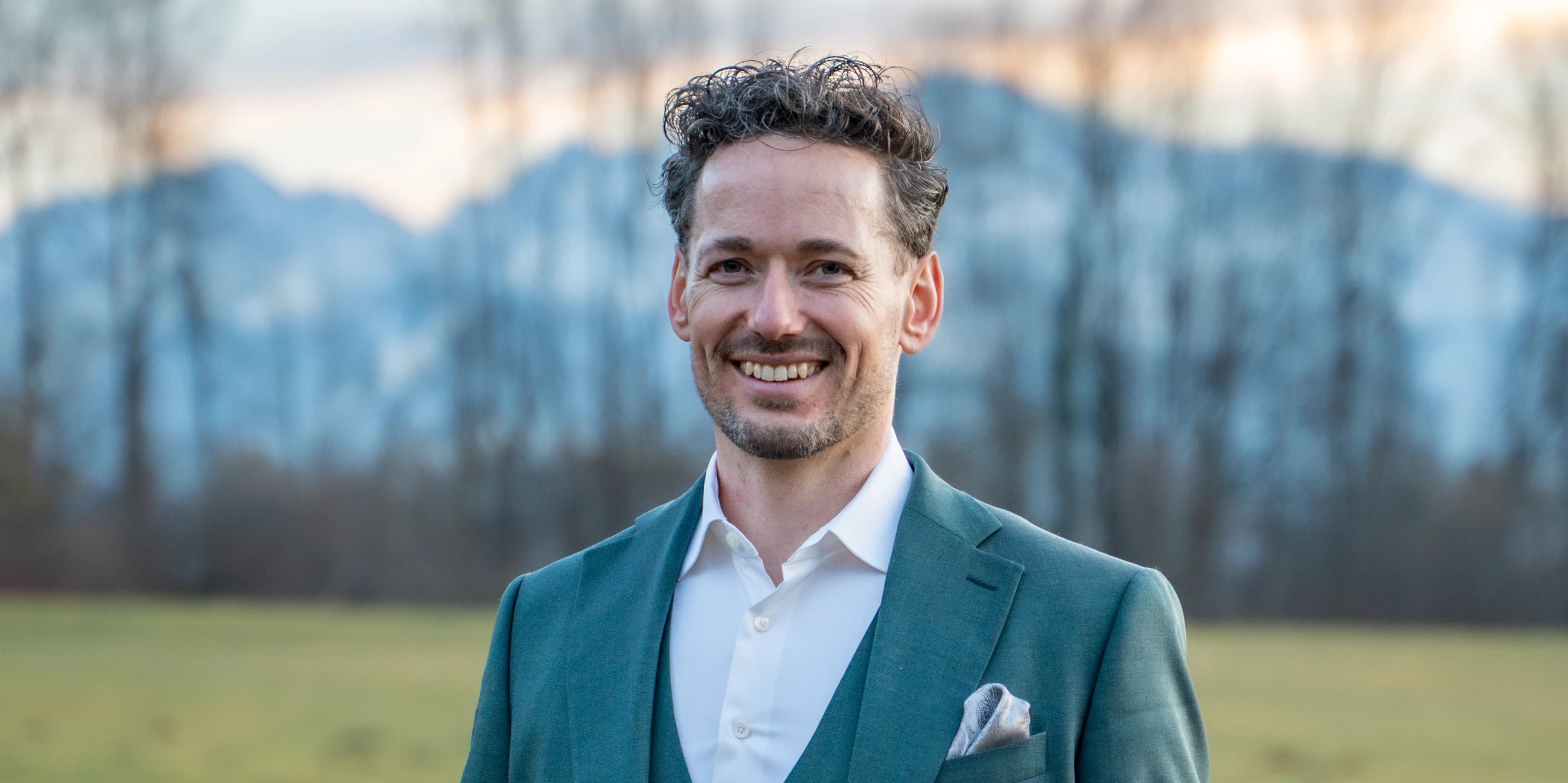 ActuQuant: Florian Brucker im grünen Anzug auf der Wiese, dahinter die Berge im Abendlicht.