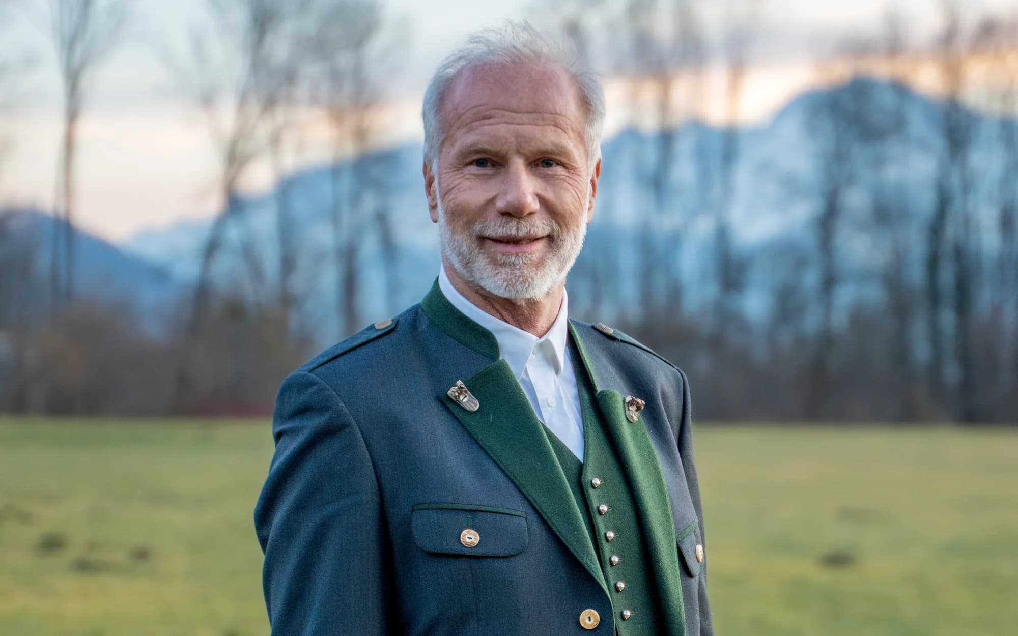 Peter Ladreiter im Trachtenanzug auf der Wiese, dahinter kahle Bäume und die Berge im Abendlicht.
