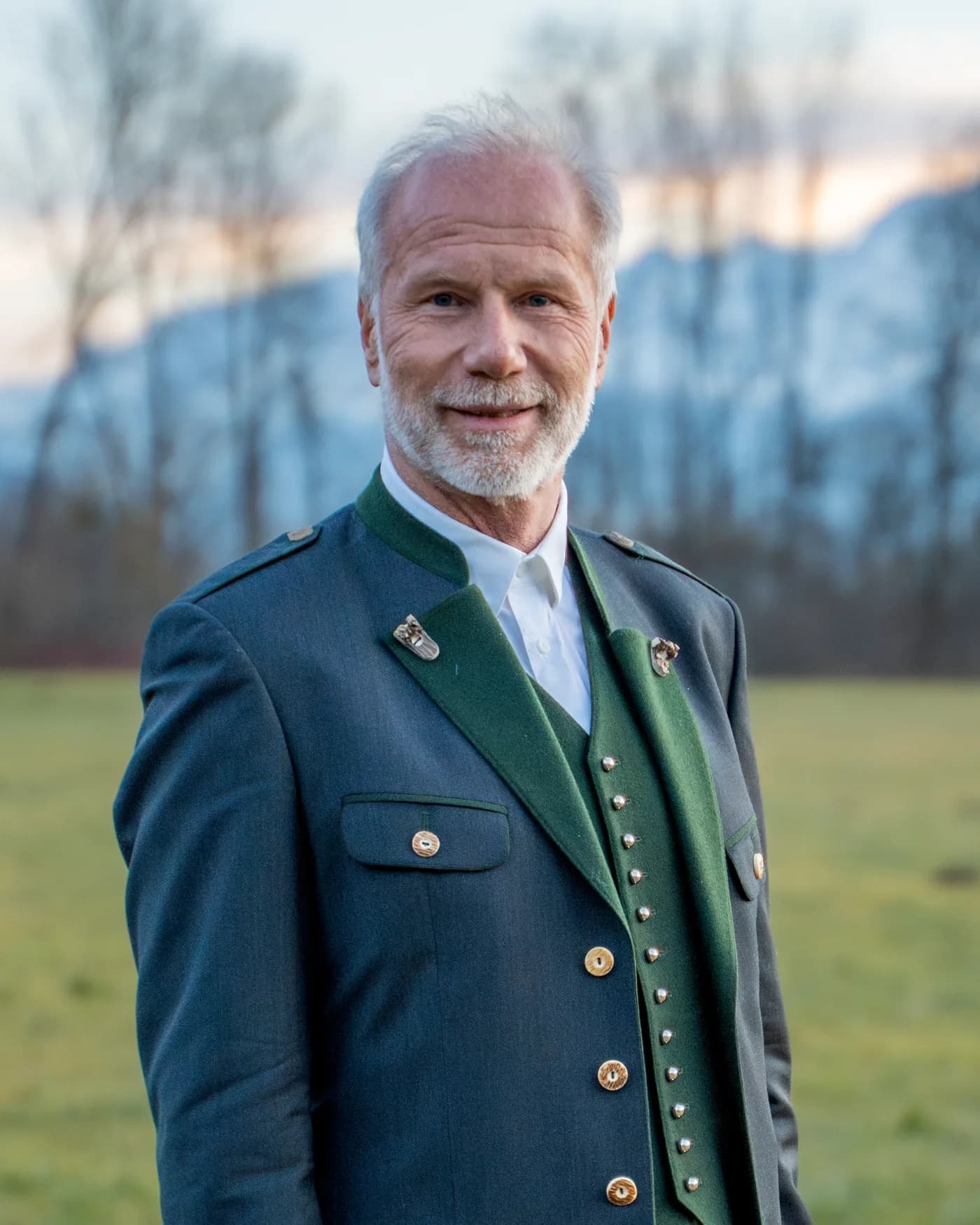 Peter Ladreiter im Trachtenanzug auf der Wiese, dahinter kahle Bäume und die Berge.
