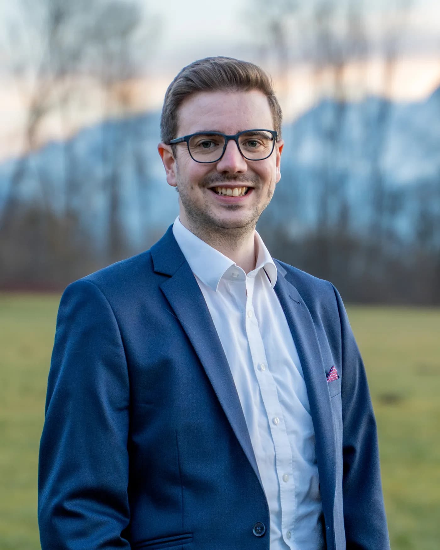 Florian Nachbagauer im blauen Anzug mit Brille auf der Wiese, dahinter die Berge.