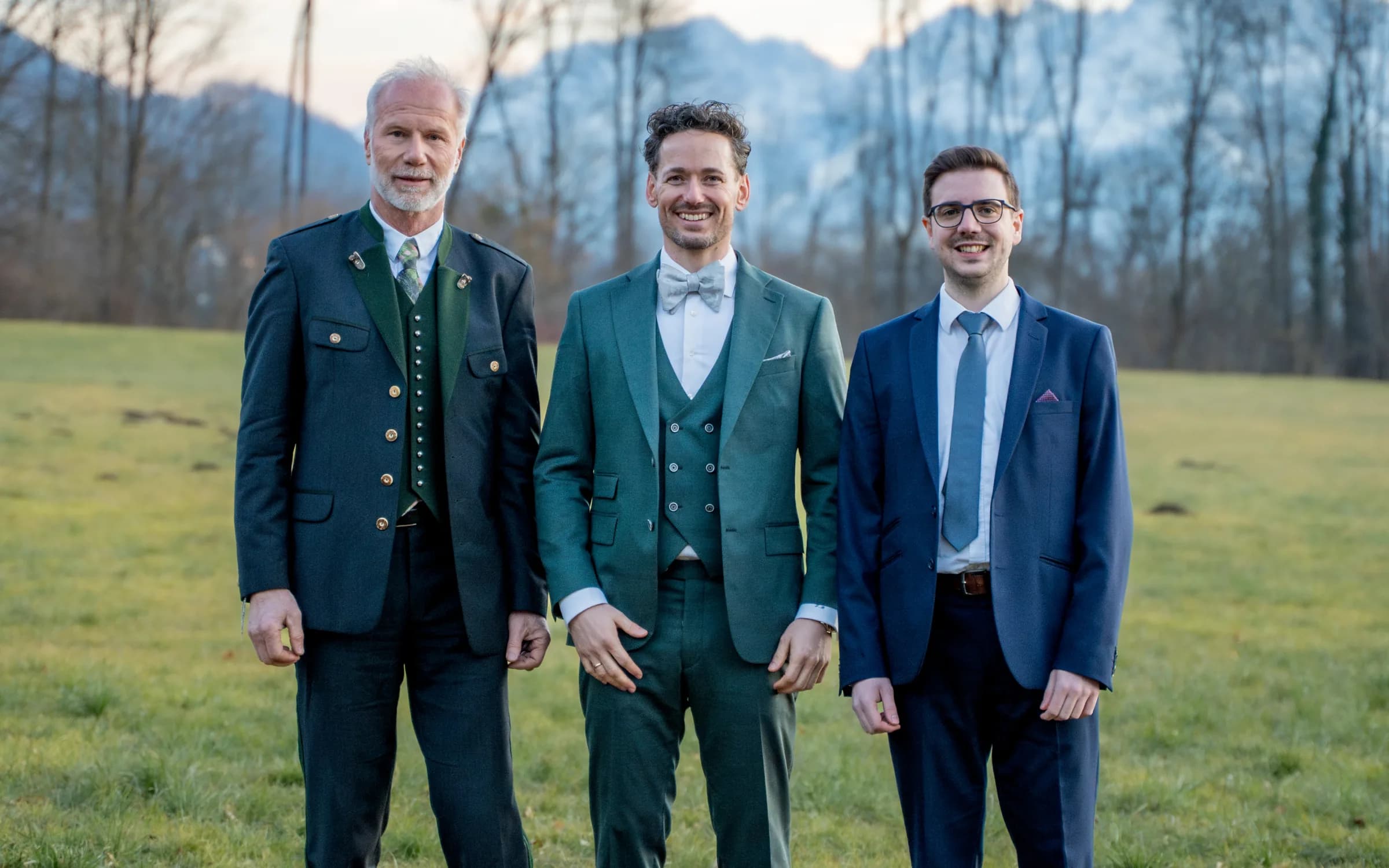 Die drei Gründer von ActuQuant stehen nebeneinander auf einer Wiese, dahinter kahle Bäume und die Berge im Abendlicht.