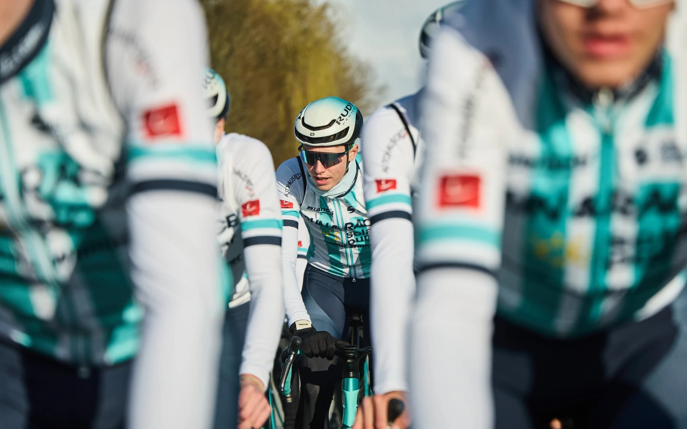 Fahrer des Teams RUN & RACE solar pur dicht nebeneinander auf der Straße, im Vordergrund unscharfe Trikotärmel, dazwischen ein Fahrer mit Helm und Sonnenbrille.