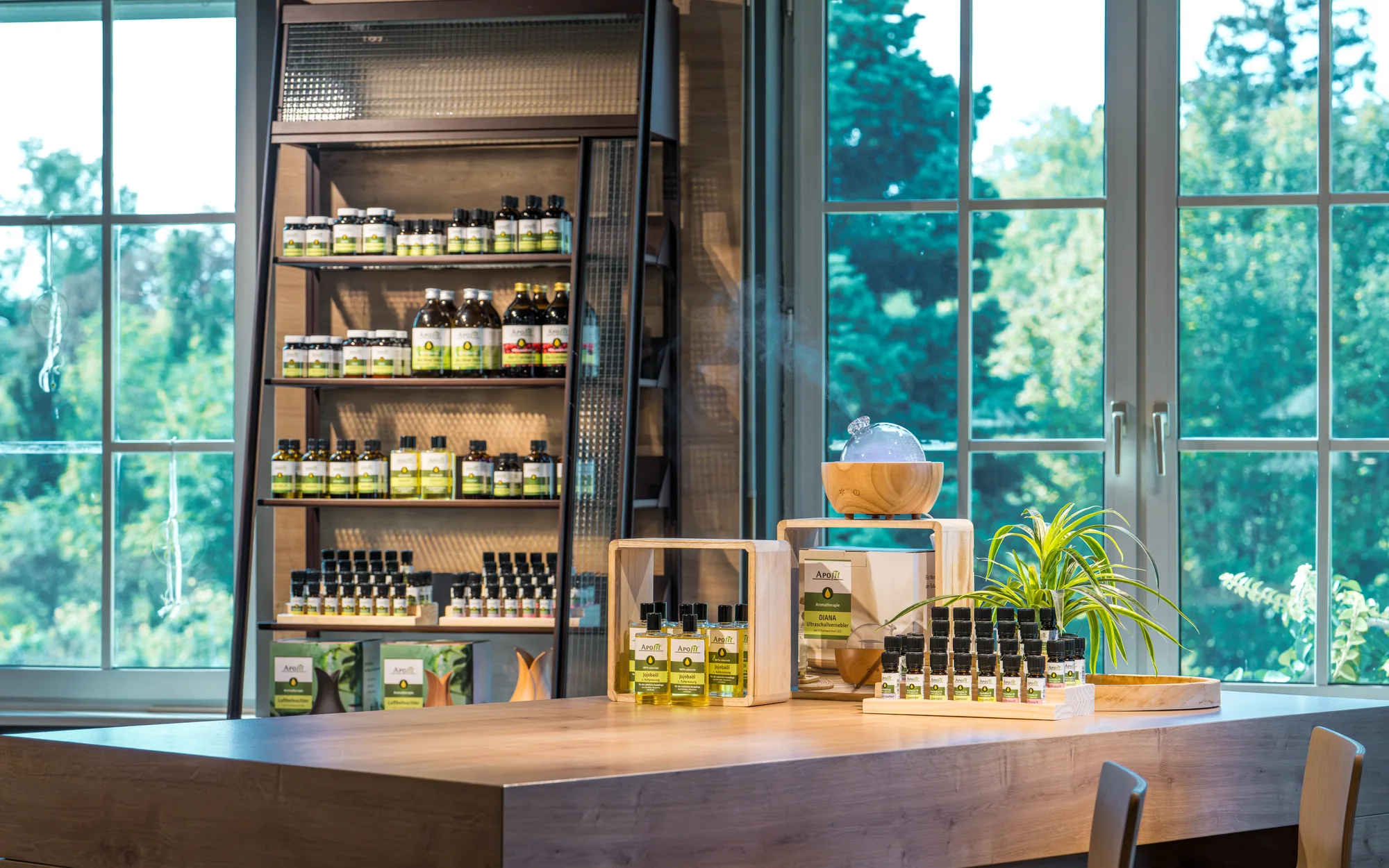 Ein Regal mit APOfit-Produkten, davor ein Schreibtisch mit ätherischen Ölen in einer Holzbox und ein Diffuser aus Holz, im Hintergrund ein großes Fenster.