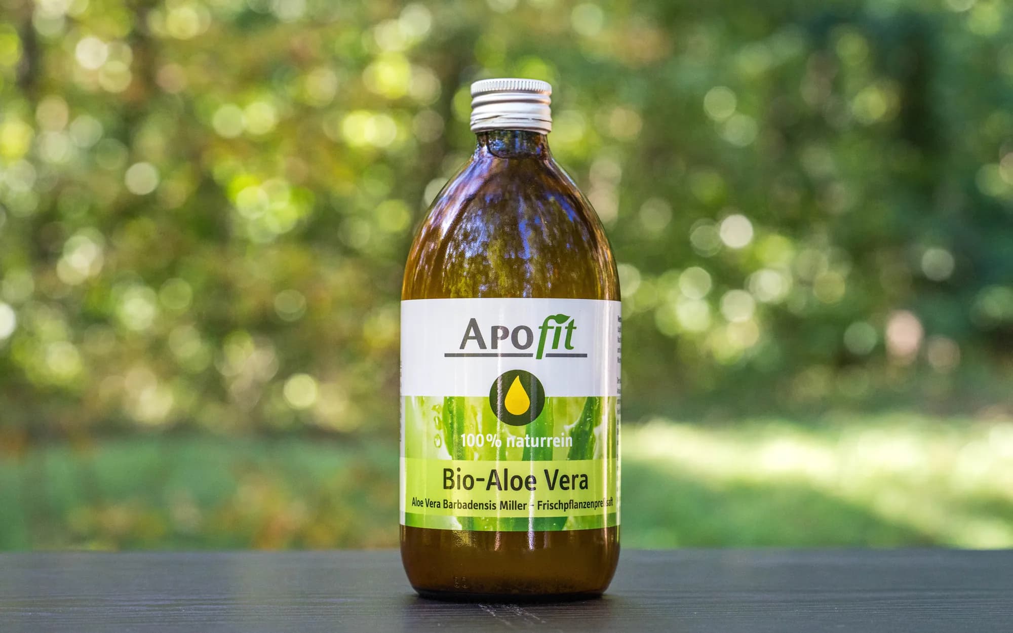 Eine Flasche APOfit Bio-Aloe-Vera-Saft steht auf einer Holzkante vor unscharfem grünem Laub.