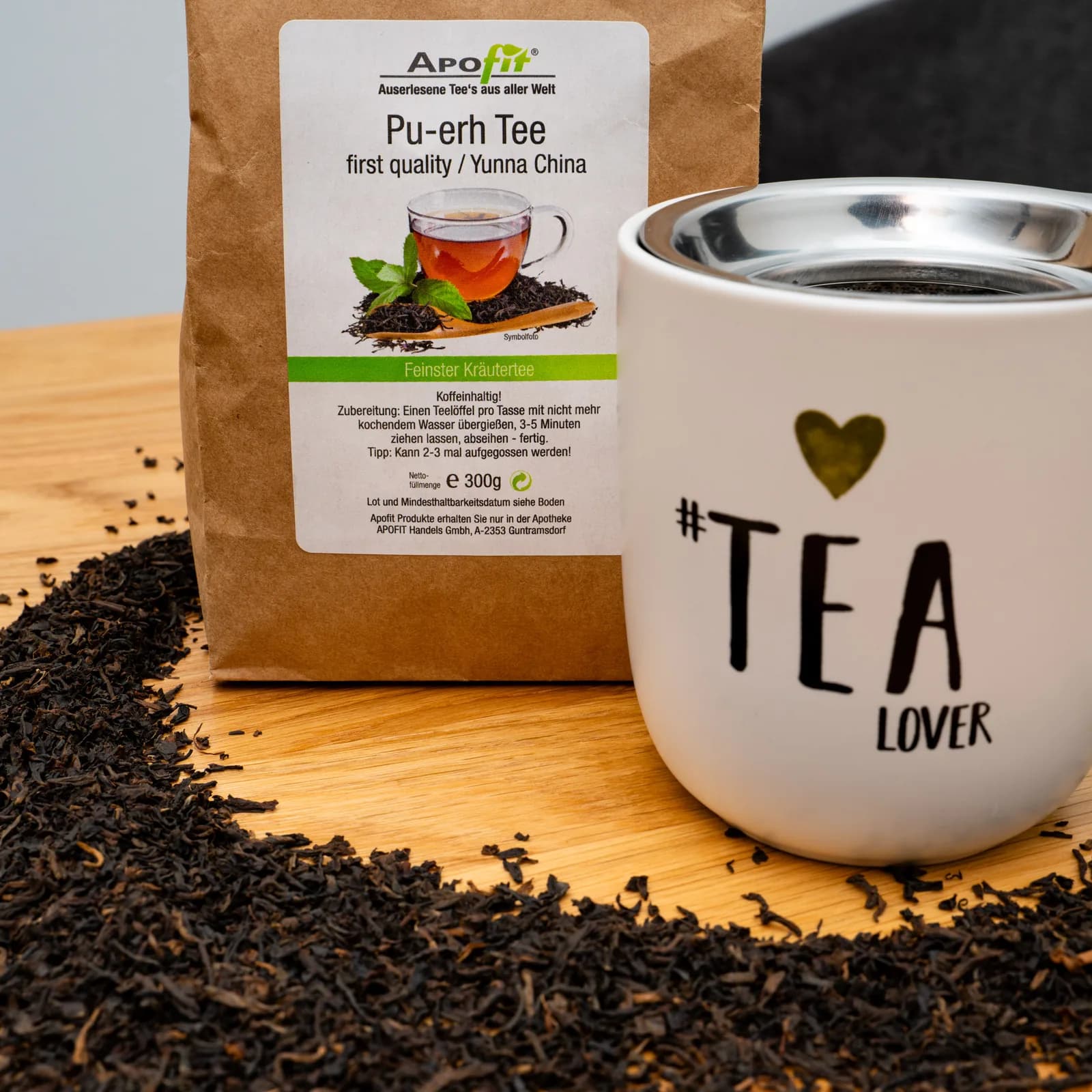 Eine Packung APOfit Pu-erh Tee steht neben einer Tasse mit der Aufschrift Tea Lover, davor ein Kranz aus losen Teeblättern auf einer Holzplatte.