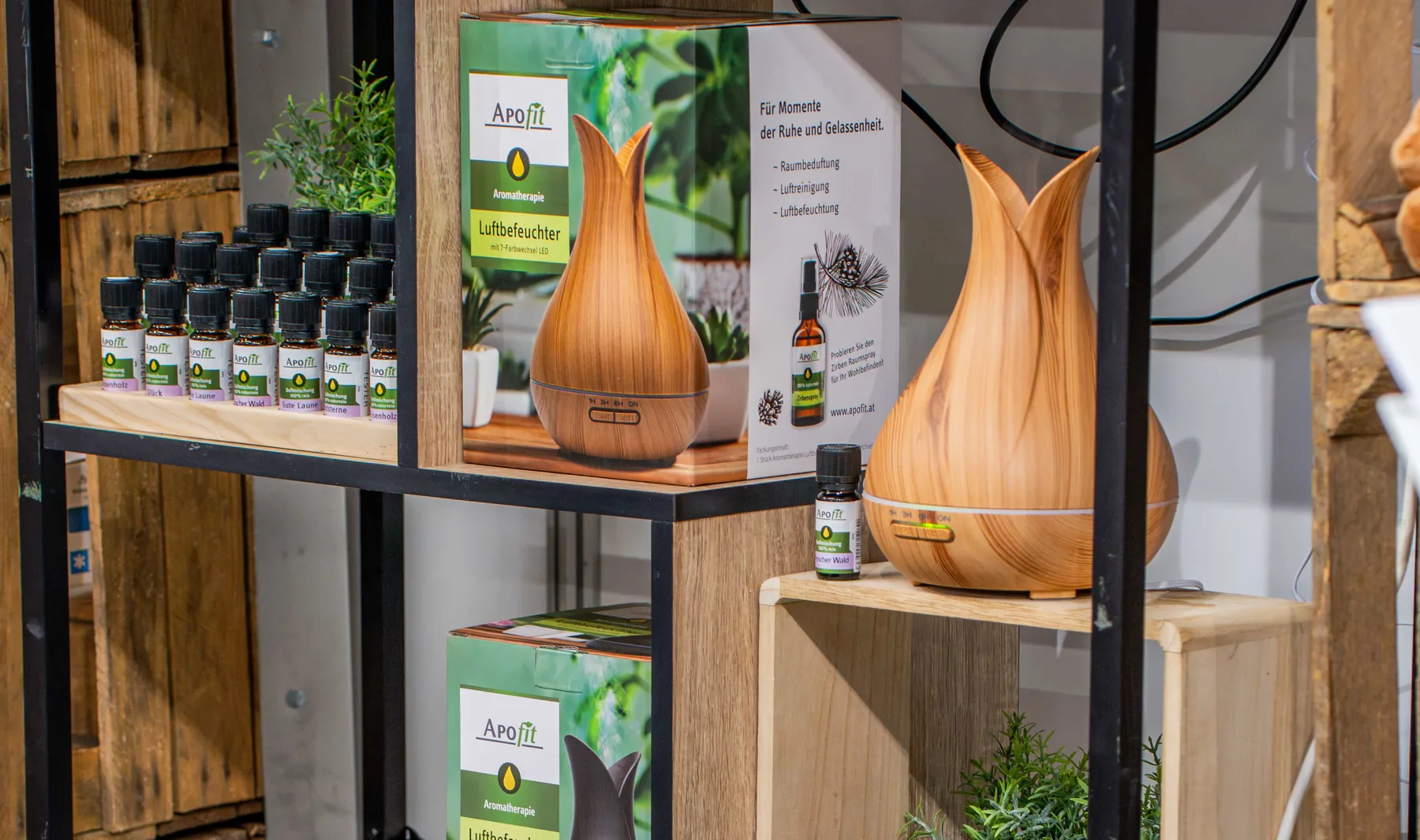Ein Regal aus Holzkisten am Messestand: die Schachtel des Luftbefeuchters mit ihrem Aufdruck, daneben der Diffuser aus Holzoptik und eine Reihe kleiner Fläschchen mit APOfit-Etikett.