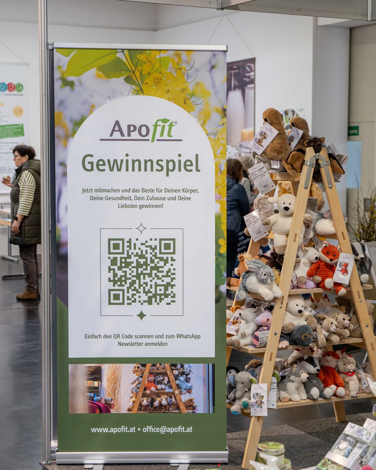 Das Roll-up zum Gewinnspiel steht in der Messehalle: Logo, Überschrift und ein großer QR-Code auf grünem Grund, daneben eine Leiter voller Plüschtiere.