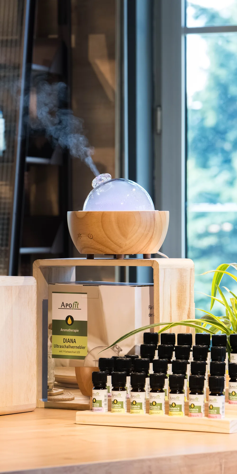 Ein Regal mit APOfit-Produkten, davor auf einem Schreibtisch eine Holzbox mit ätherischen Ölen und ein Diffuser aus Holz, aus dem Dampf steigt, dahinter ein großes Fenster.