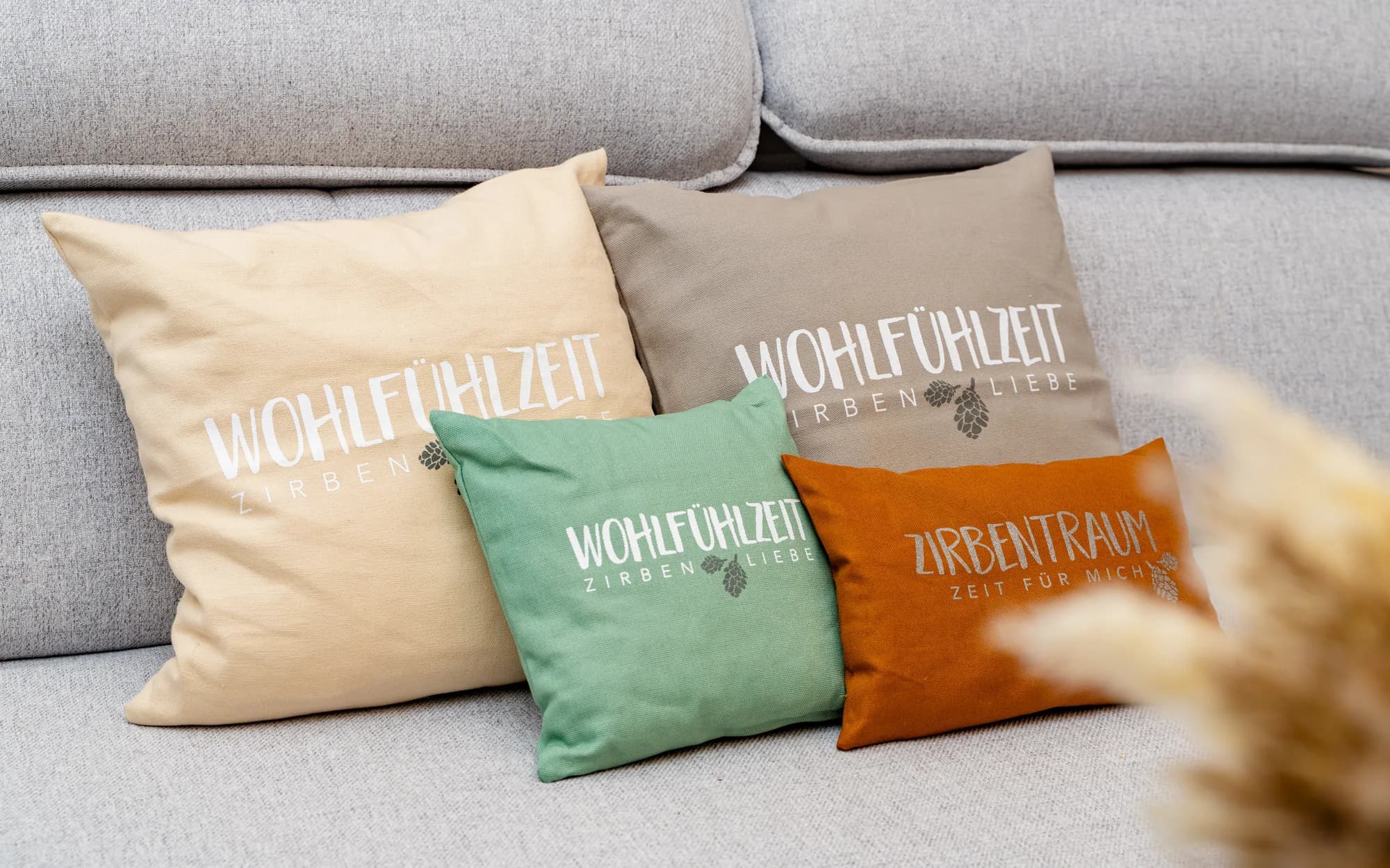 Vier Kissen mit den Aufschriften Wohlfühlzeit, Zirbenliebe und Zirbentraum auf einem grauen Sofa, links und rechts Trockengräser.