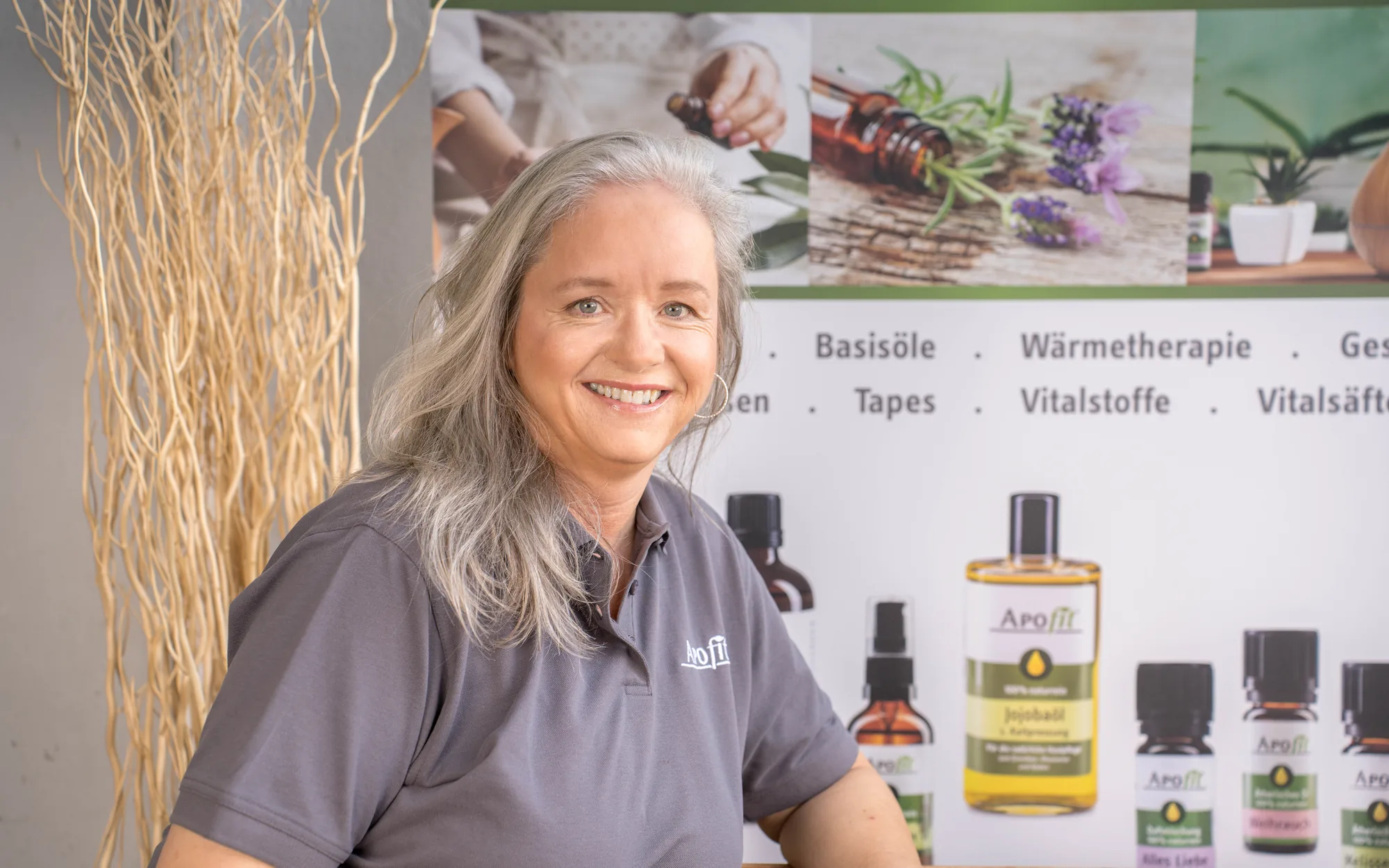 Eine Mitarbeiterin von APOfit in grauem Poloshirt sitzt lächelnd vor einem Banner mit dem Sortiment: Basisöle, Wärmetherapie, Tapes, Vitalstoffe, Vitalsäfte.