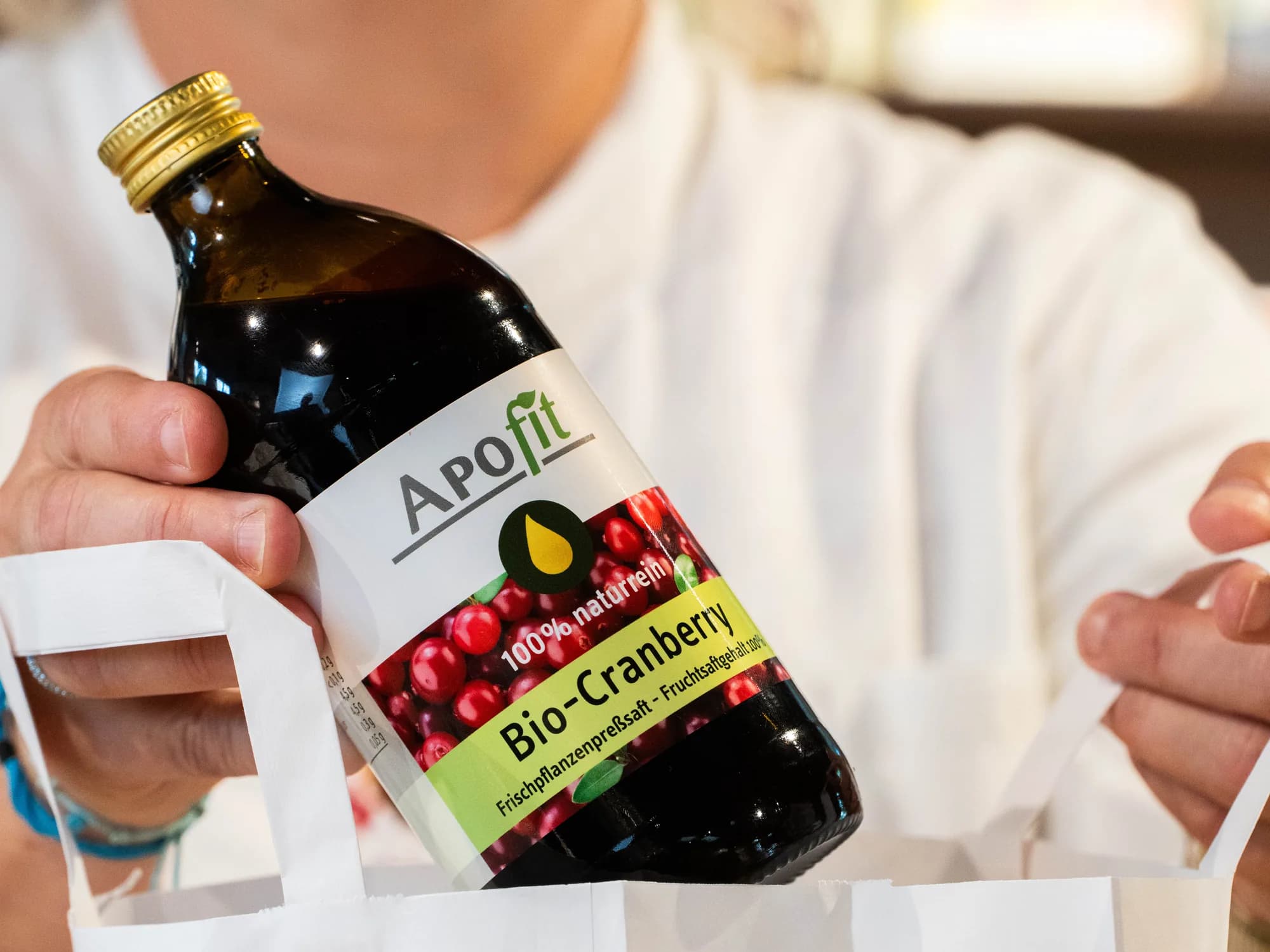 Eine Apothekerin im weißen Mantel legt eine Flasche APOfit Bio-Cranberry-Saft in eine weiße Papiertasche mit Blattmotiv.