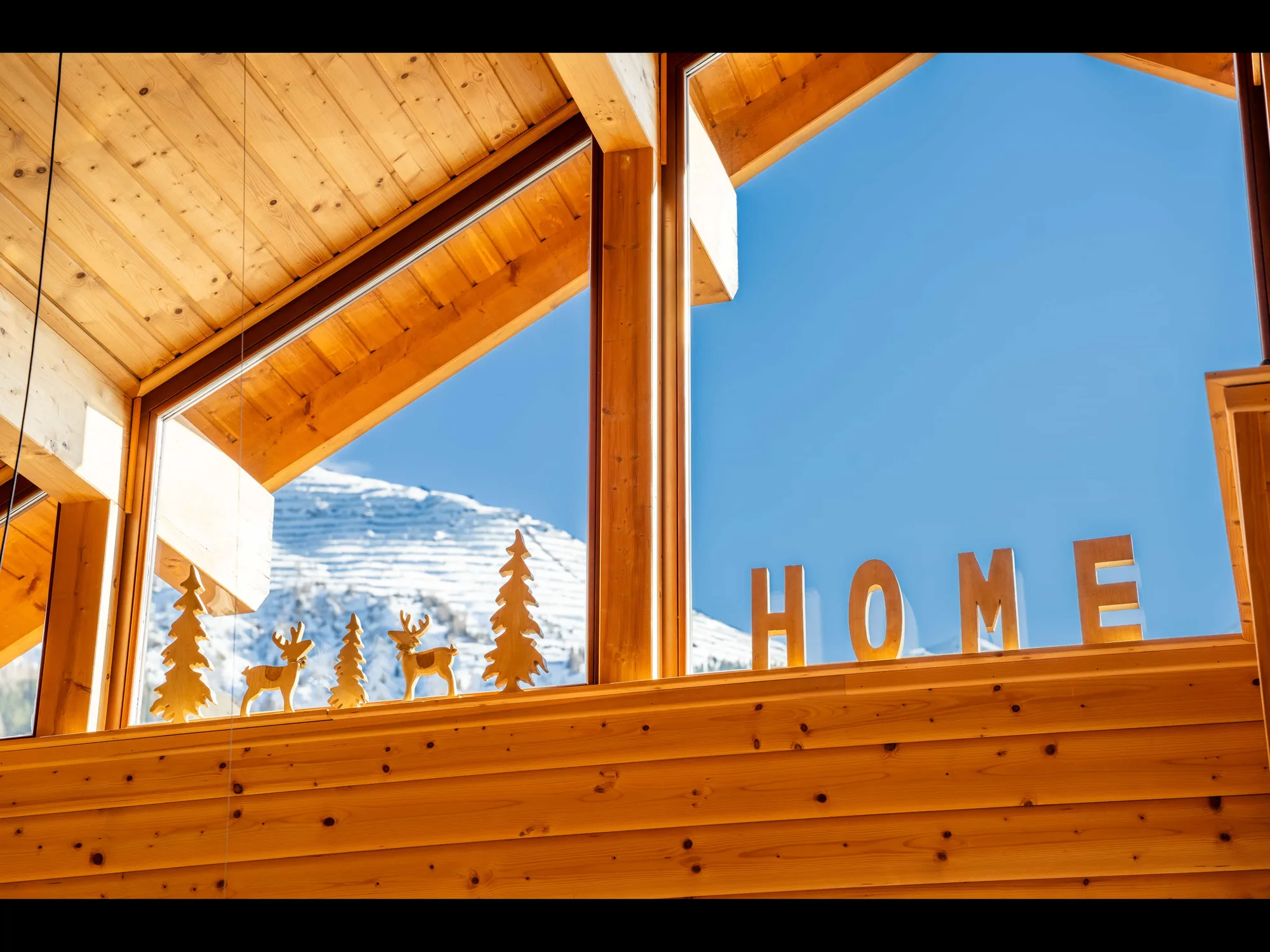 Das Giebelfenster der Arlberg Kandahar Lodge von innen: der Schriftzug HOME aus Holzbuchstaben auf der Fensterbank, dahinter der verschneite Berg.