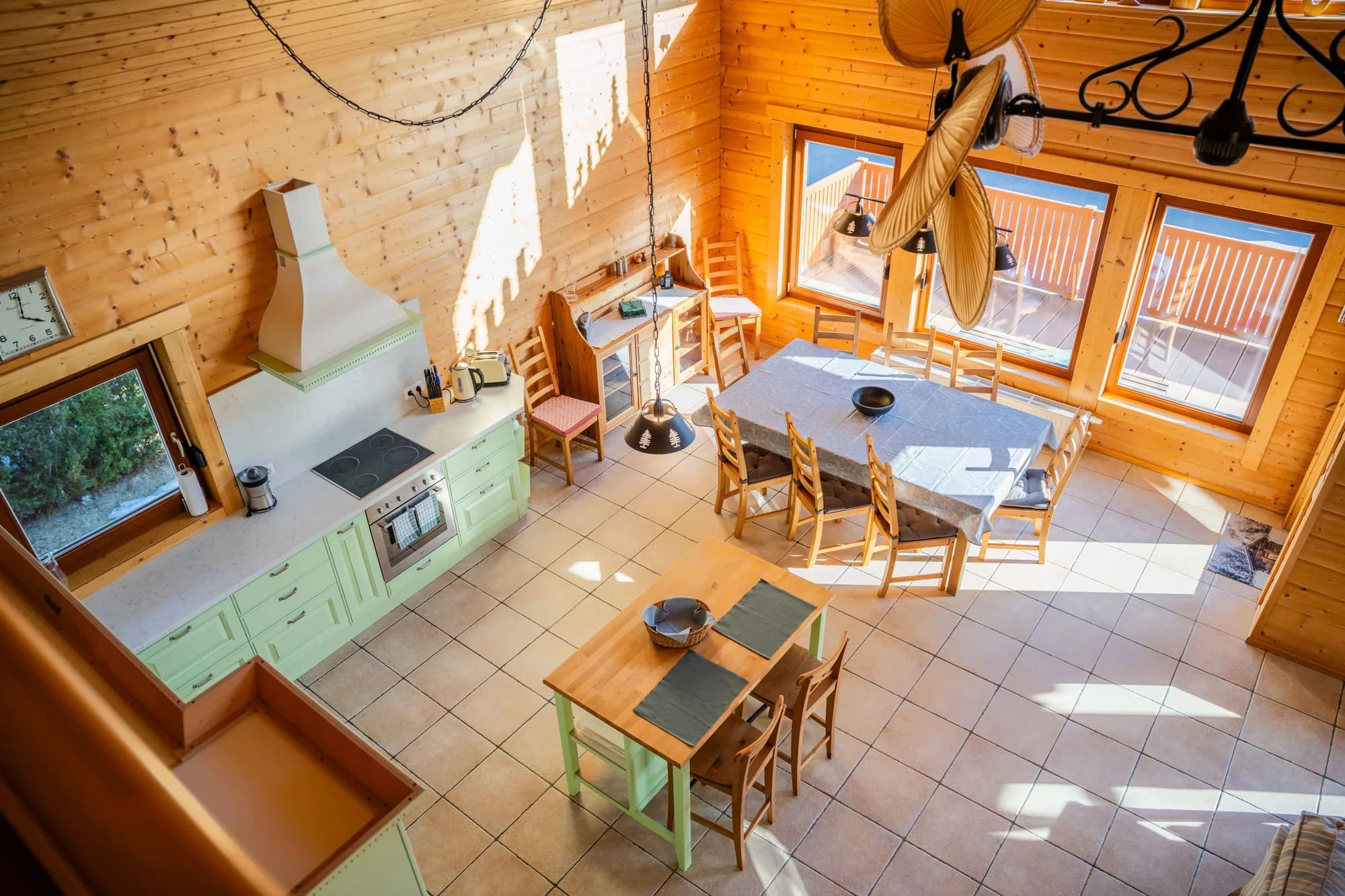 Die Küche der Arlberg Kandahar Lodge: mintgrüne Fronten, eine Kochinsel mit Holztisch und dahinter der Essplatz an den Fenstern.