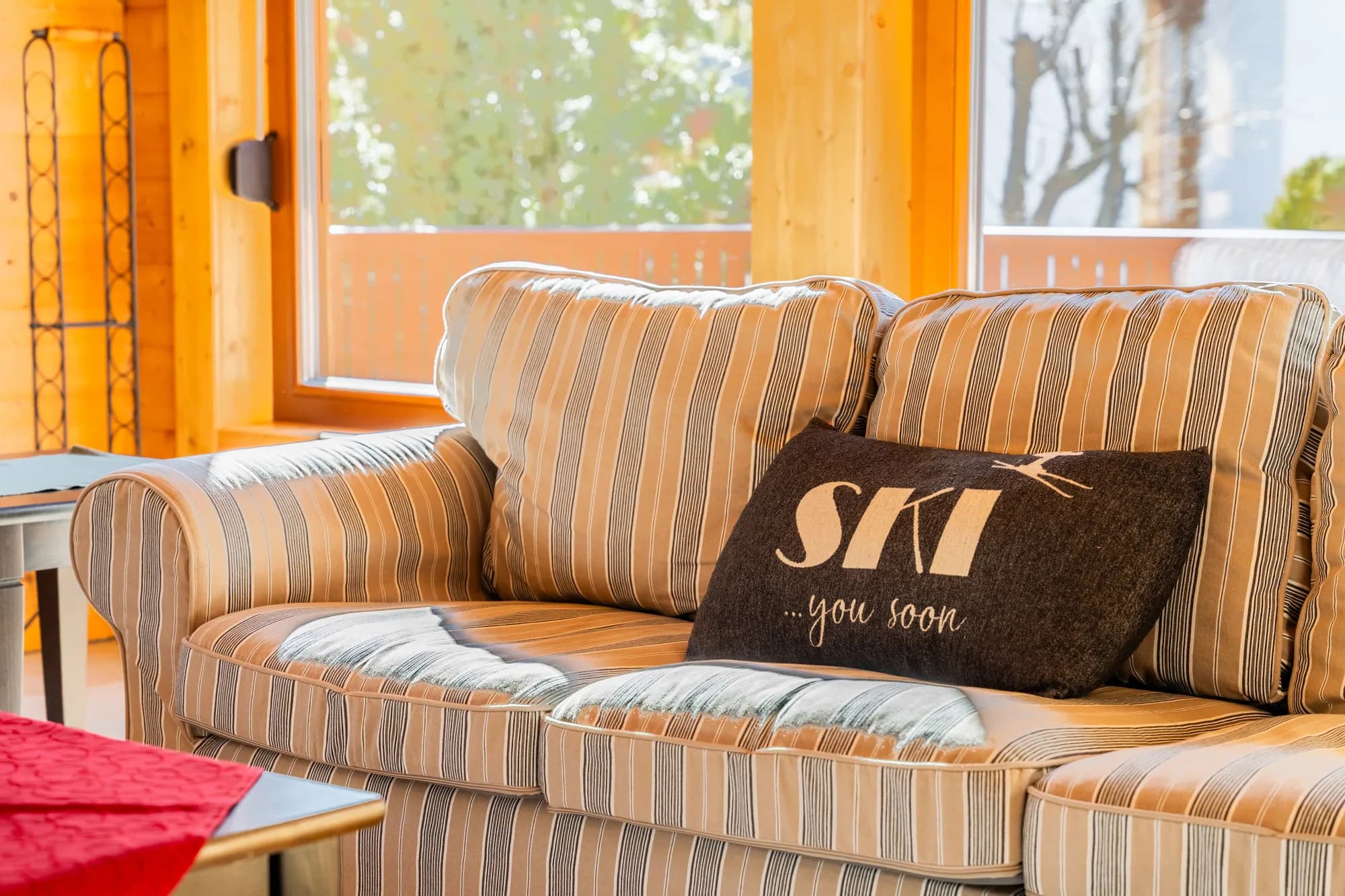 Ein Sofa im Wohnraum der Arlberg Kandahar Lodge, darauf ein Kissen mit der Aufschrift SKI you soon, dahinter das Fenster mit Blick auf die Bäume.