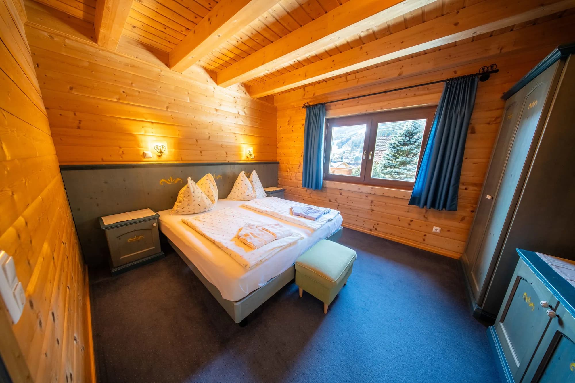 Das blaue Schlafzimmer der Arlberg Kandahar Lodge: ein Doppelbett unter der Holzdecke, blauer Teppich, Fenster mit Blick auf die Tannen.