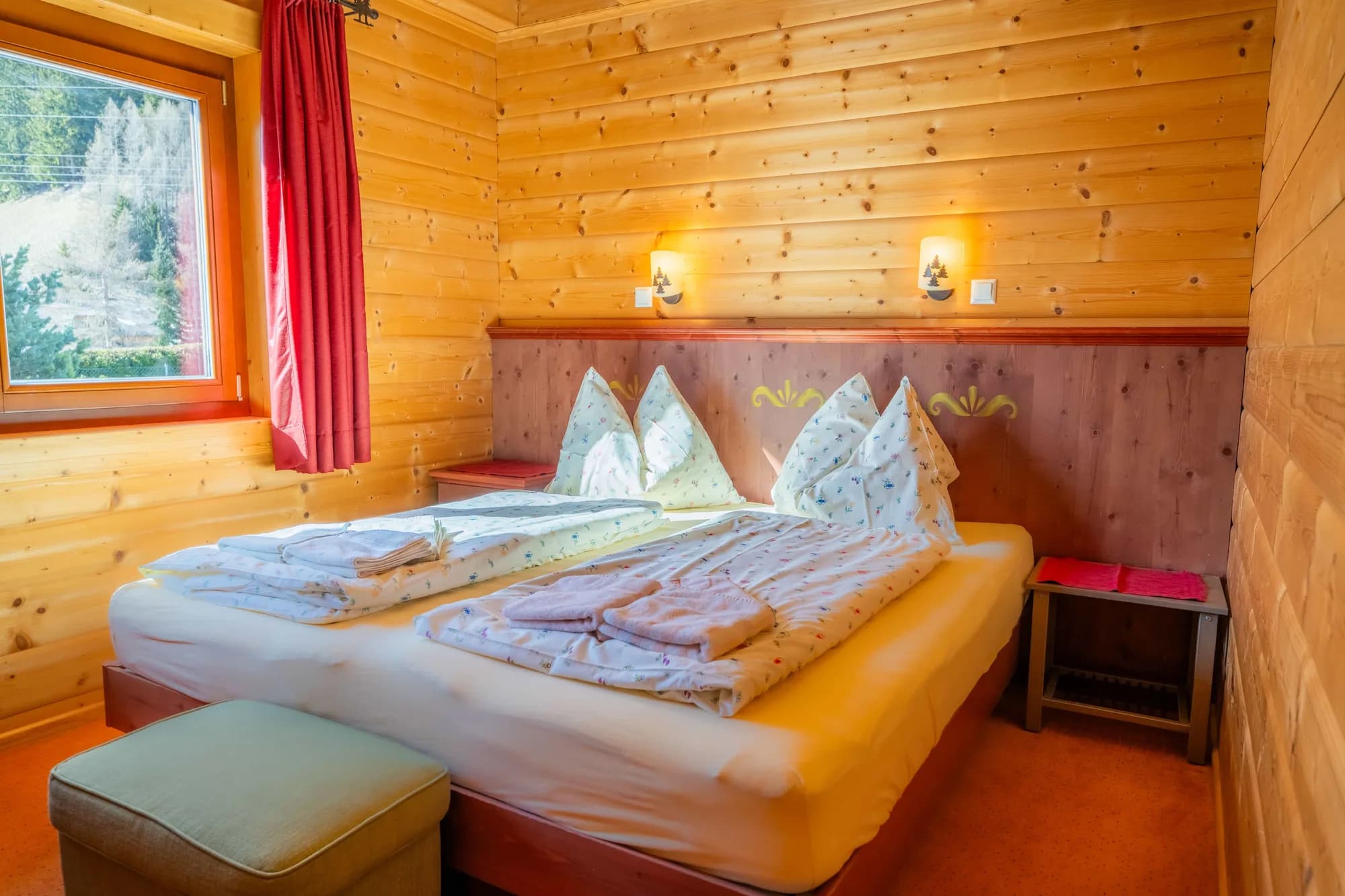 Das rote Schlafzimmer der Arlberg Kandahar Lodge: ein Doppelbett zwischen zwei Nachtlampen, davor eine gepolsterte Bank, daneben das Fenster.