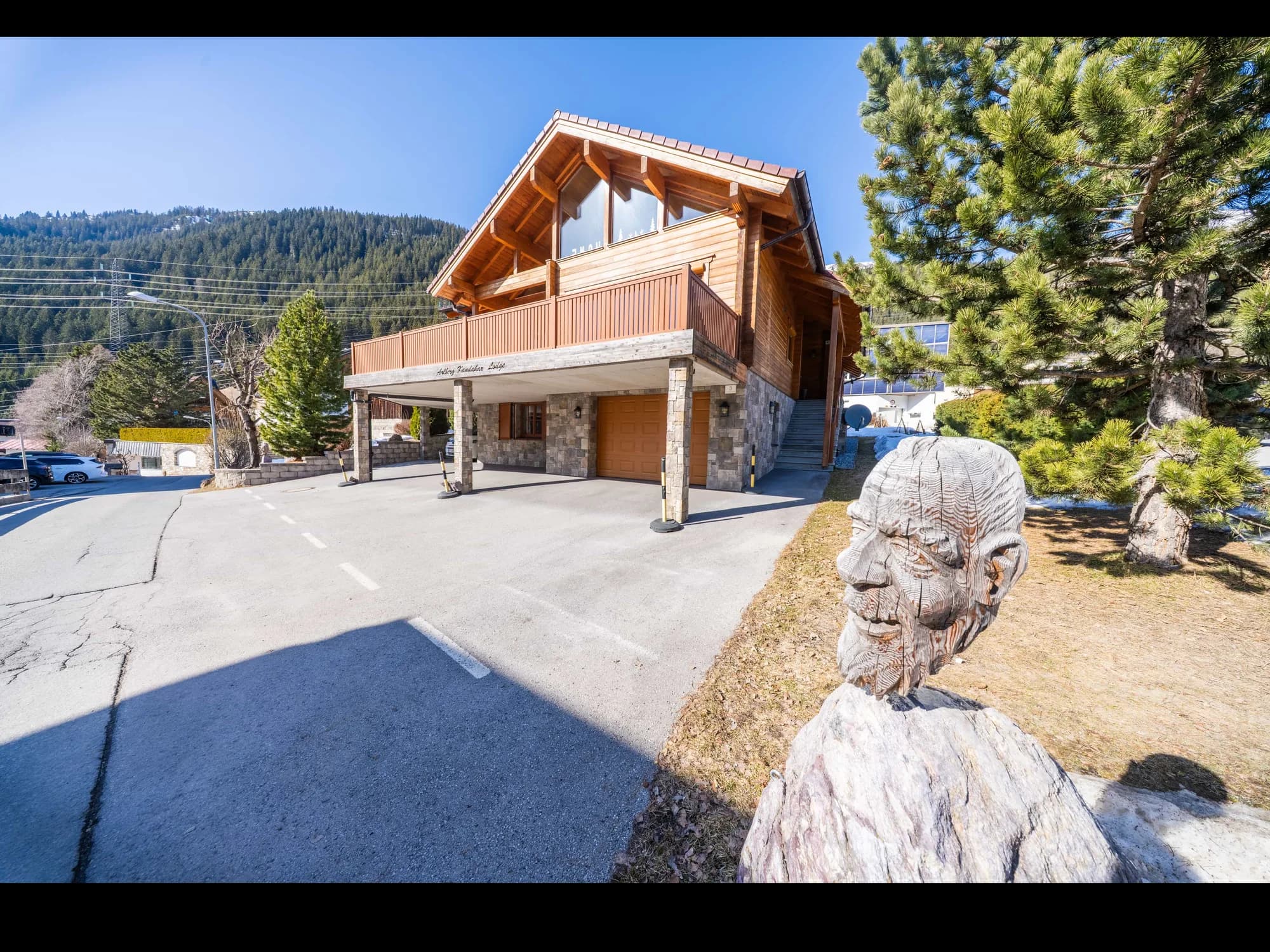 Die Arlberg Kandahar Lodge von der Straße aus: das Holzhaus mit überdachtem Vorplatz und Balkon, dahinter der bewaldete Hang.