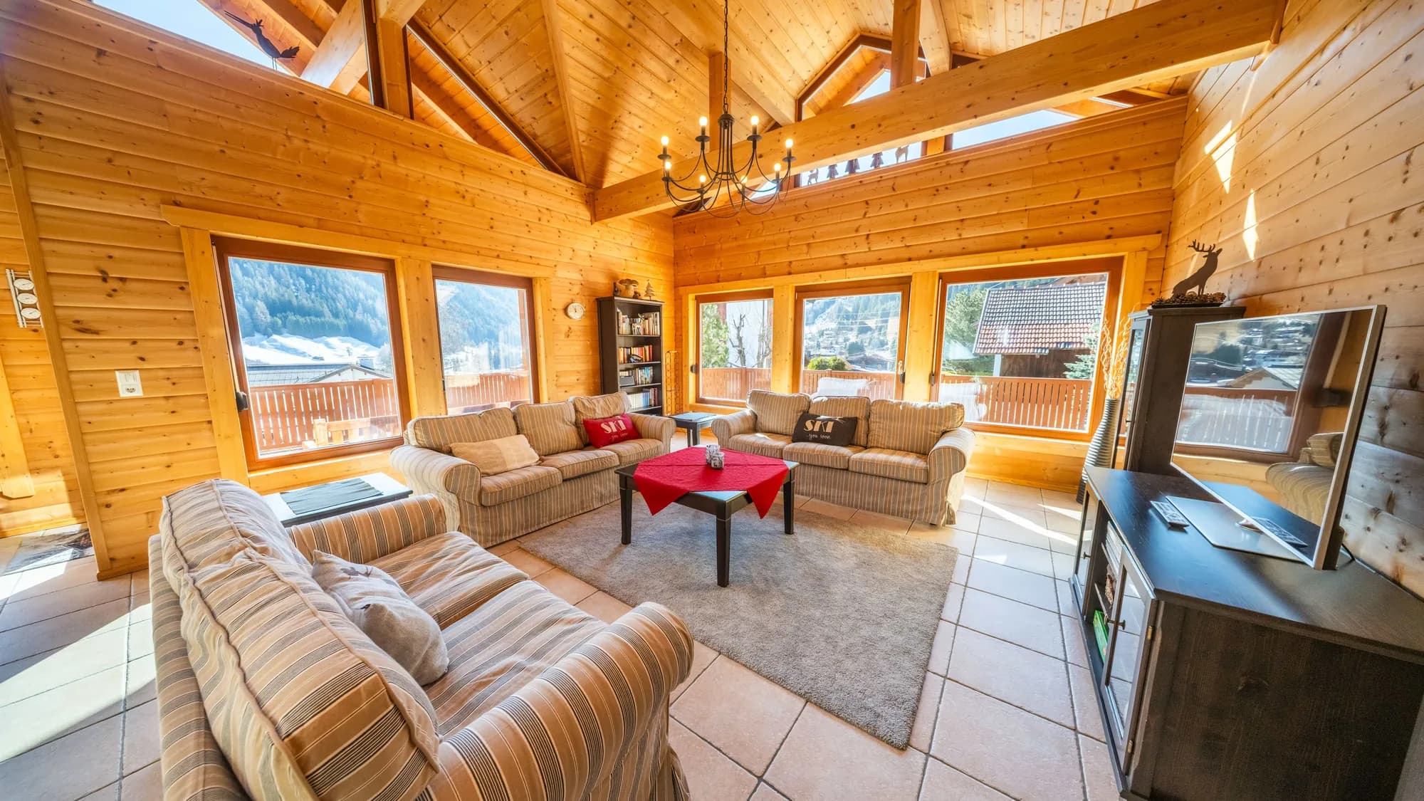 Der Wohnraum der Arlberg Kandahar Lodge: heller Holzgiebel über zwei Sofagarnituren, ein roter Tisch in der Mitte, dahinter drei Fenster mit Blick auf die Berge.