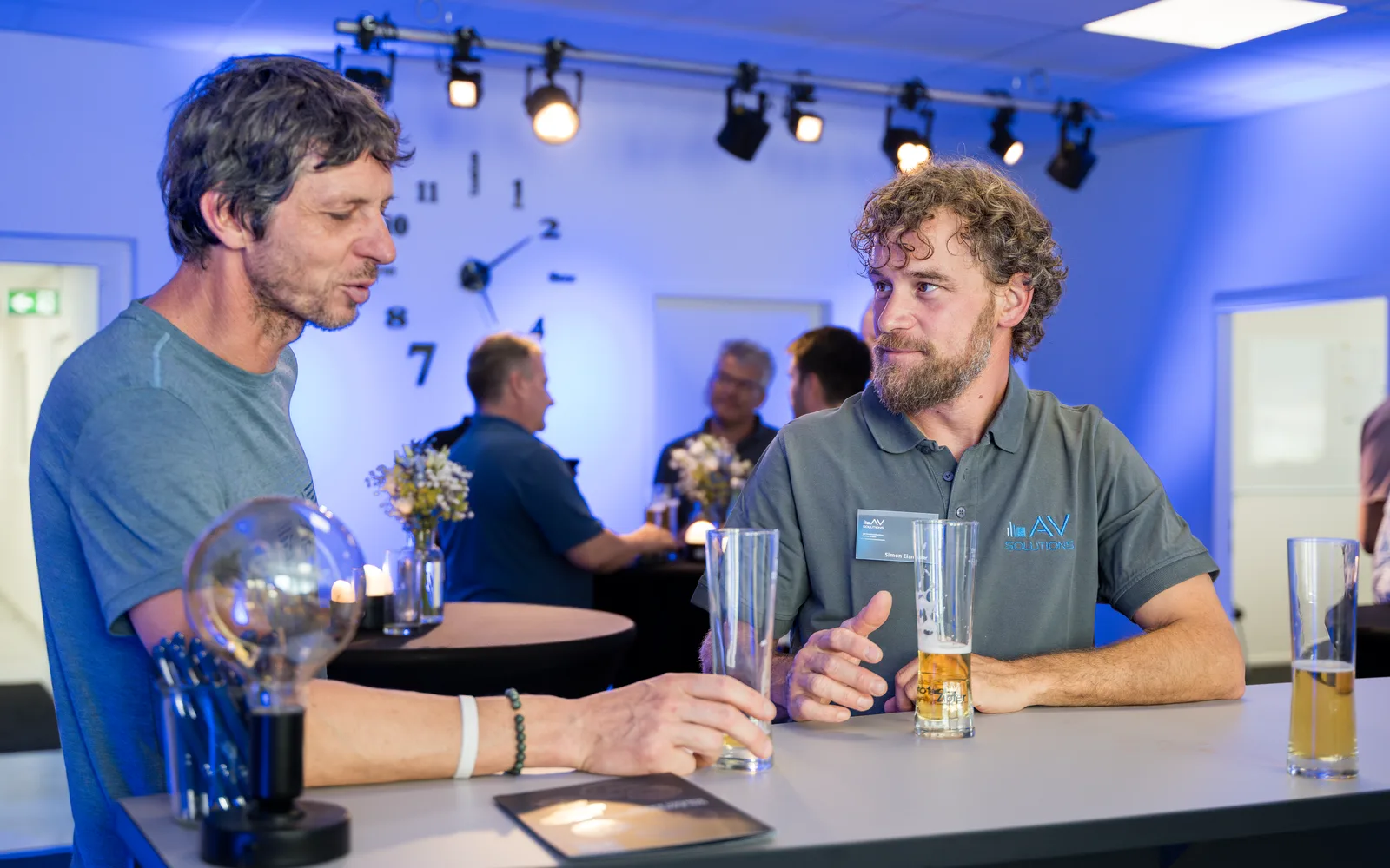 Zwei Männer stehen im blau ausgeleuchteten Raum an einem hohen Tisch mit Biergläsern und unterhalten sich, im Hintergrund weitere Gäste und eine große Wanduhr.
