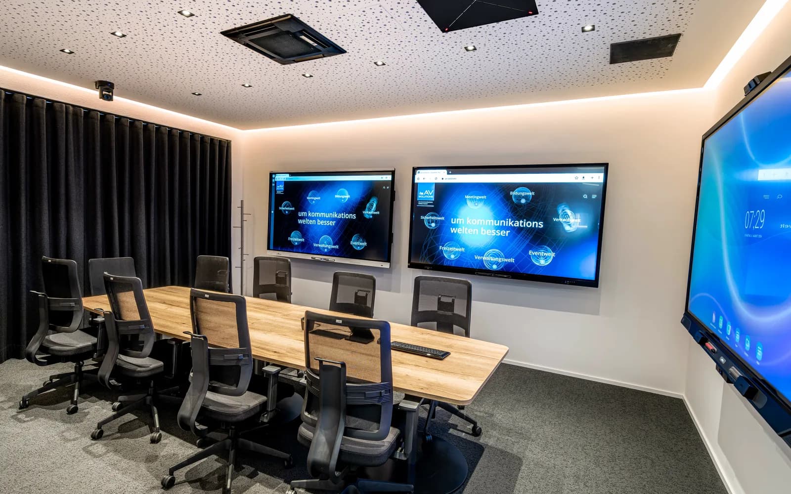 Ein Besprechungsraum von AVsolutions mit langem Holztisch und schwarzen Stühlen, an der Wand zwei Bildschirme mit dem Satz um kommunikationswelten besser und rechts ein großes interaktives Display.