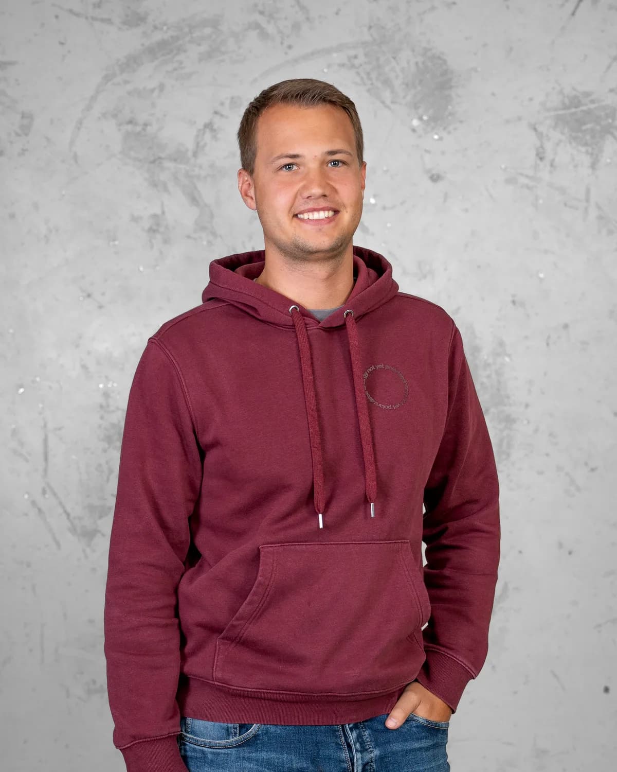 Ein junger Mann mit kurzem blondem Haar trägt einen weinroten Kapuzenpullover und steht mit einer Hand in der Hosentasche vor einer grauen Betonwand.