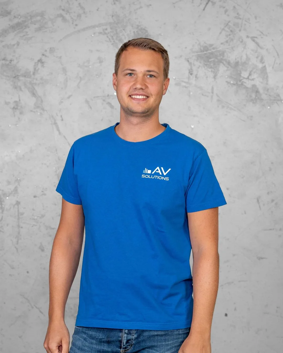 Ein junger Mann mit kurzem blondem Haar im blauen Firmen-T-Shirt steht mit hängenden Armen vor einer grauen Betonwand.