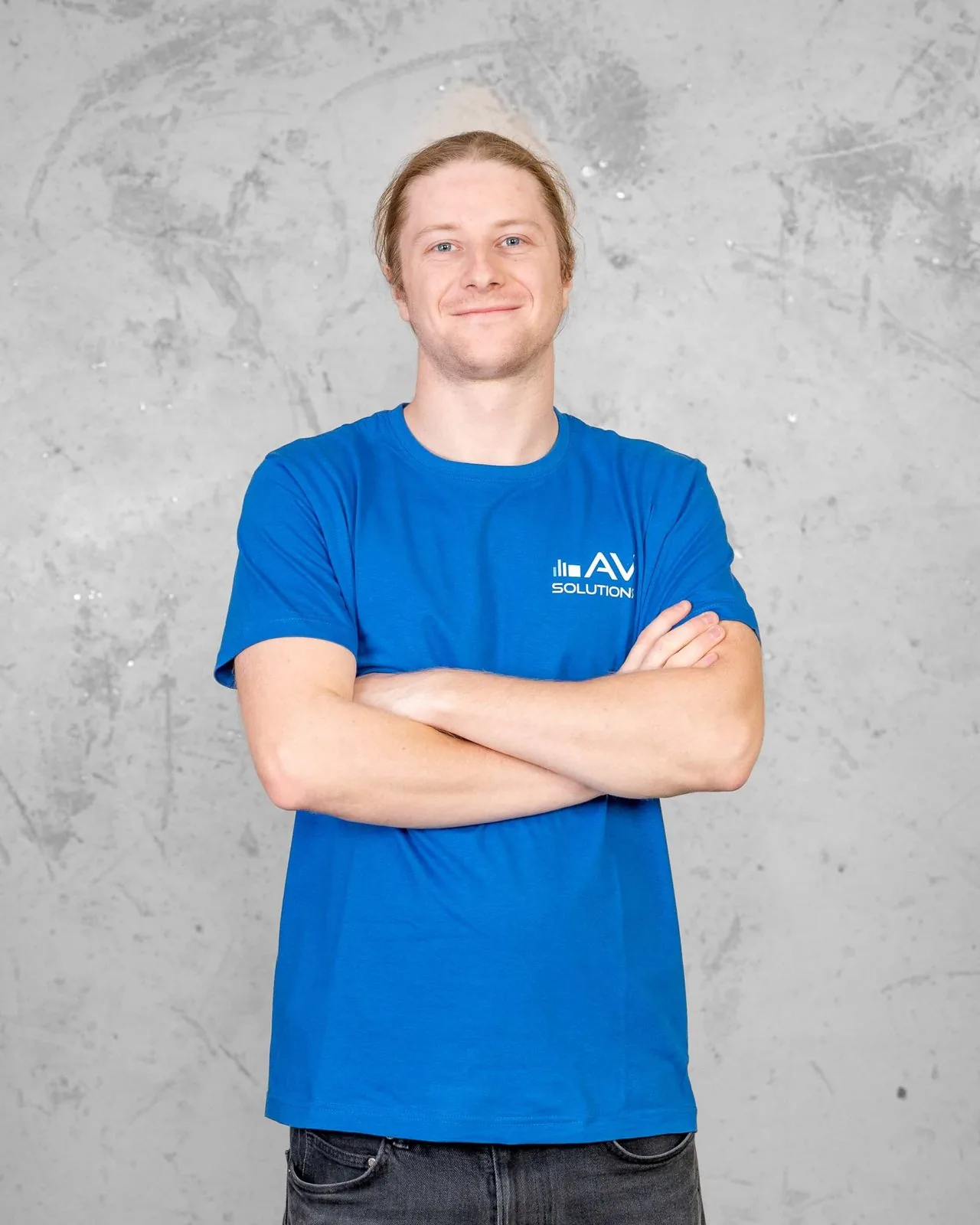 Ein junger Mann mit zurückgebundenem blondem Haar im blauen Firmen-T-Shirt steht mit verschränkten Armen vor einer grauen Betonwand.