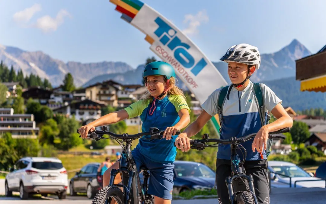 Zwei Kinder stehen mit E-Mountainbikes unter einem Wegweiser mit der Aufschrift Gschwandtkopf, dahinter der Ort und das Karwendel.