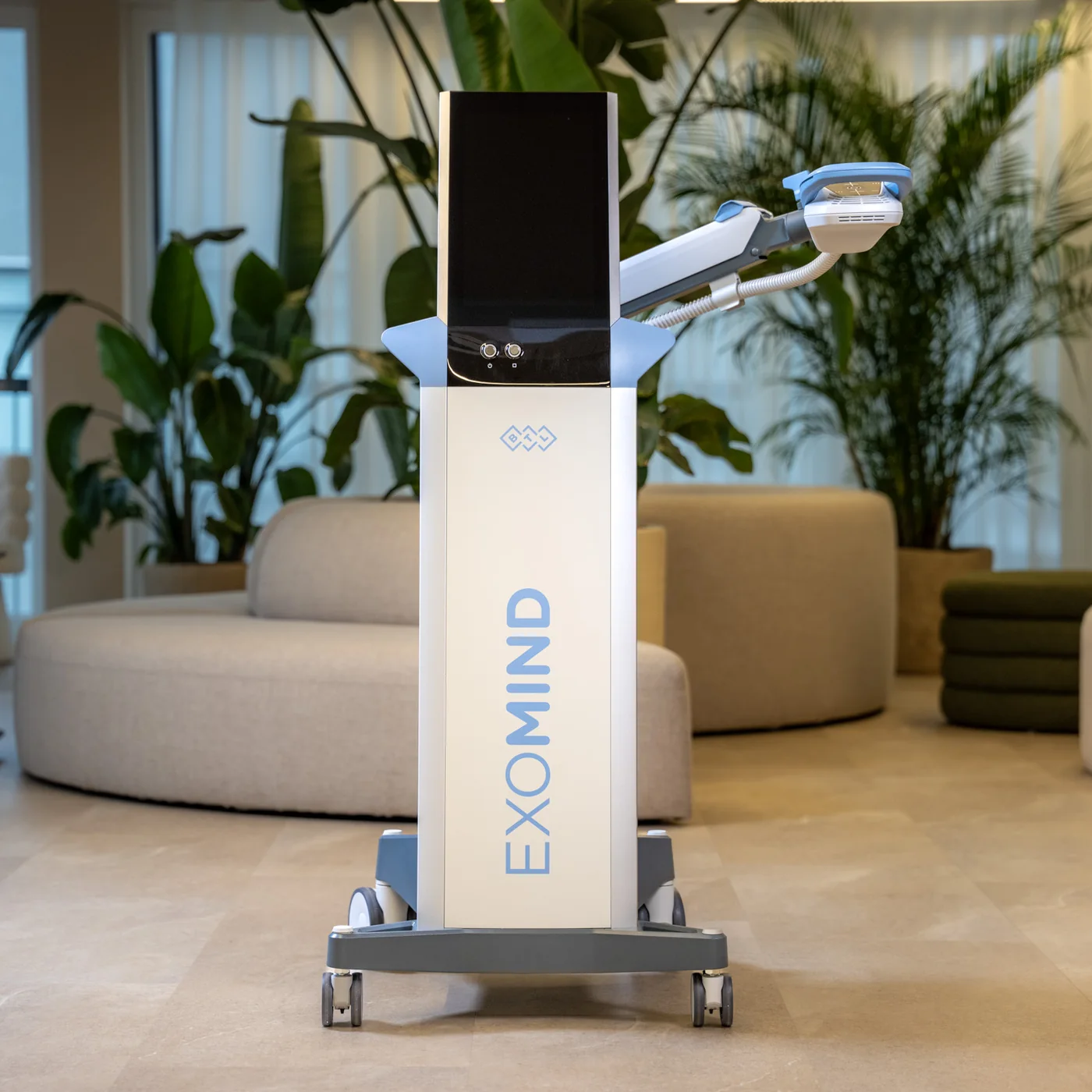 Ein EXOMIND-Gerät mit aufgeklapptem Bildschirm und Applikatorarm steht in einem hellen Raum mit Pflanzen und runden Sitzmöbeln.