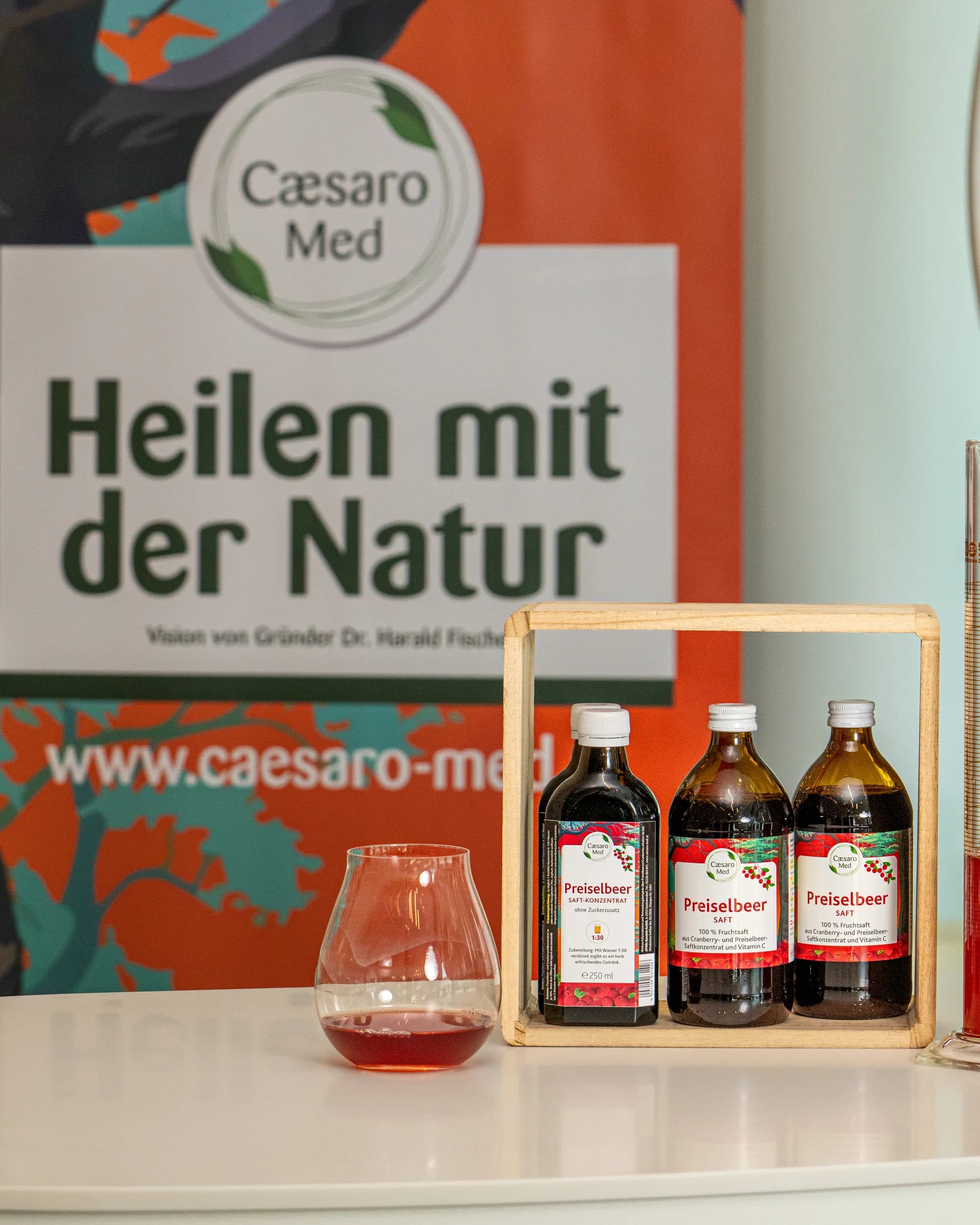 Caesaro-Med-Produkte auf dem Sichtwahltisch einer Apotheke: drei Preiselbeer-Flaschen und drei Dosen PreiselCaps in Holzrahmen, dahinter ein Rollup mit der Zeile „Heilen mit der Natur“.