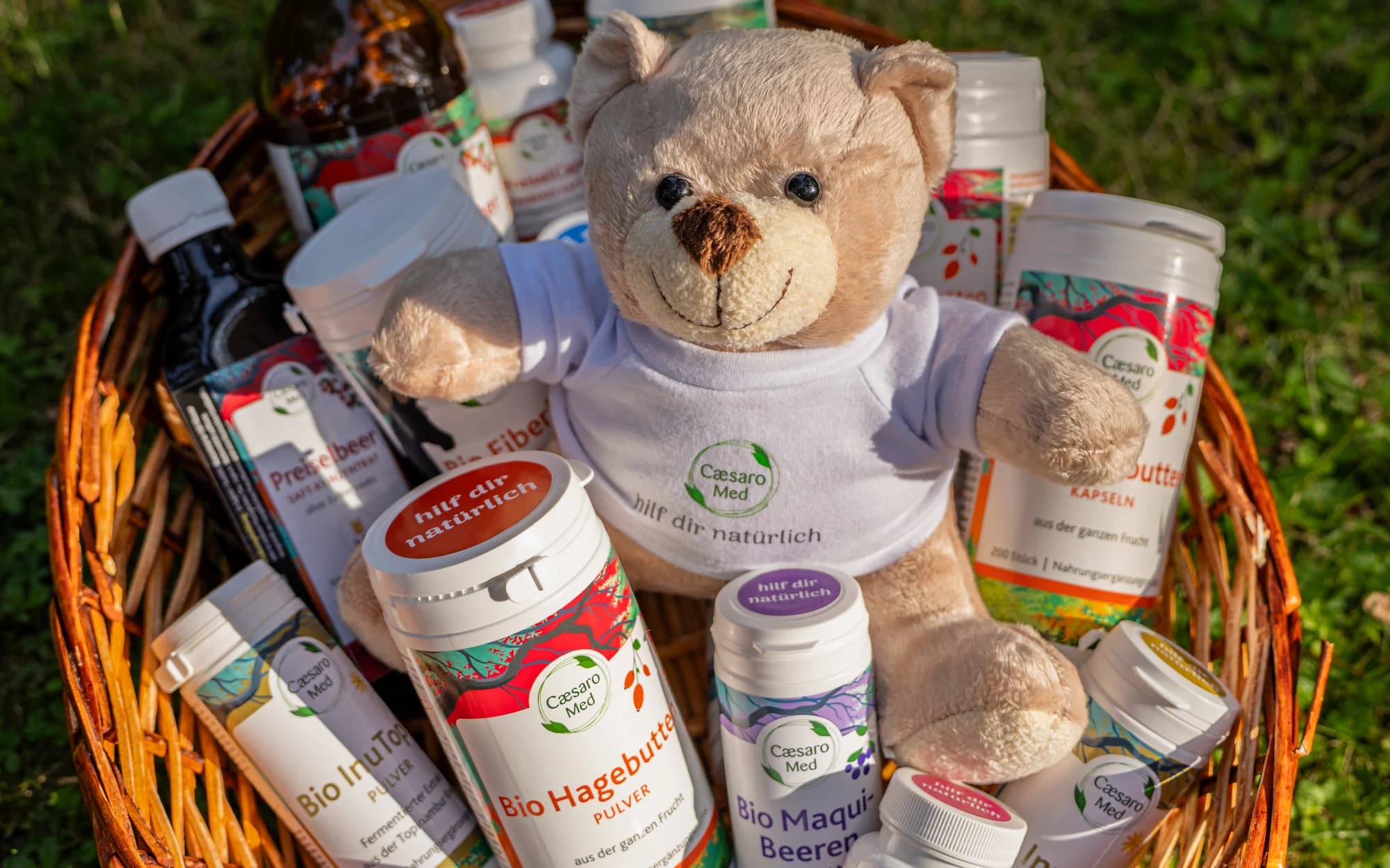 Ein Weidenkorb voller Produkte von Caesaro Med, Dosen und Flaschen dicht an dicht, dazwischen ein Plüschteddy mit dem Schriftzug hilf dir natürlich.