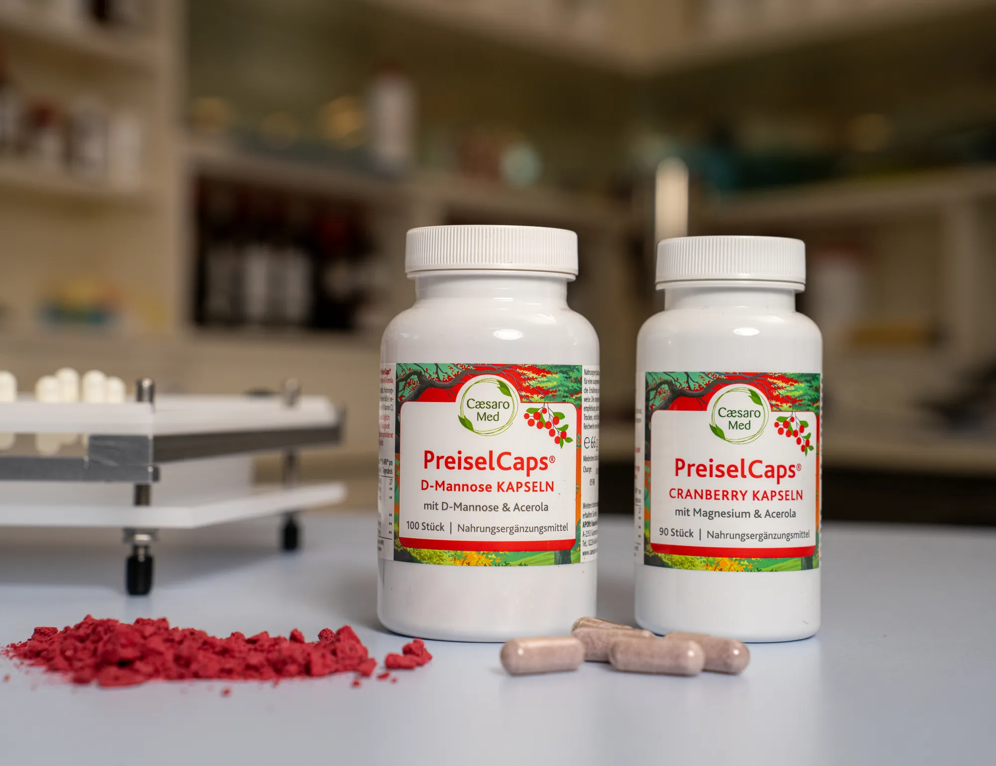 Zwei Dosen PreiselCaps stehen auf dem hellen Tresen einer Apotheke, davor liegen gefriergetrocknete Cranberrystücke und drei Kapseln, dahinter unscharf die Regale.