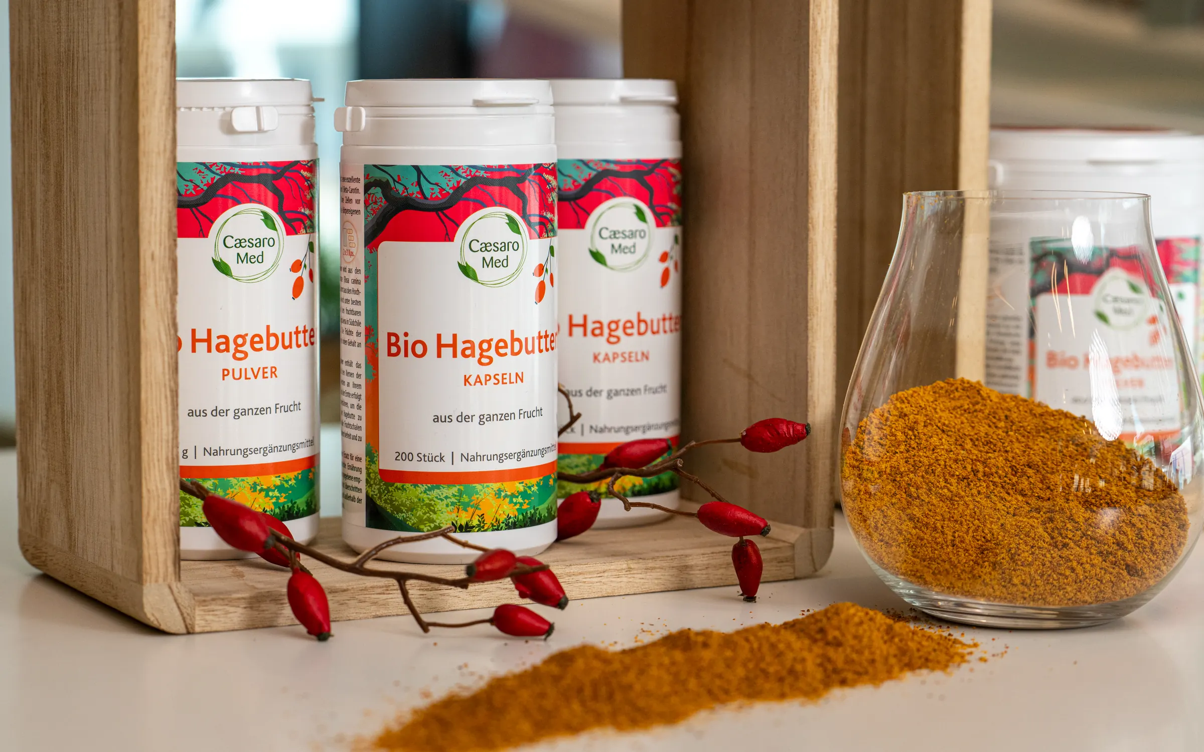 Drei Dosen Bio Hagebutten stehen in einer Holzkiste auf dem hellen Tresen einer Apotheke, davor liegt ein Zweig mit getrockneten Hagebutten und eine Spur orangefarbenes Pulver, rechts daneben ein Glas voll Pulver.
