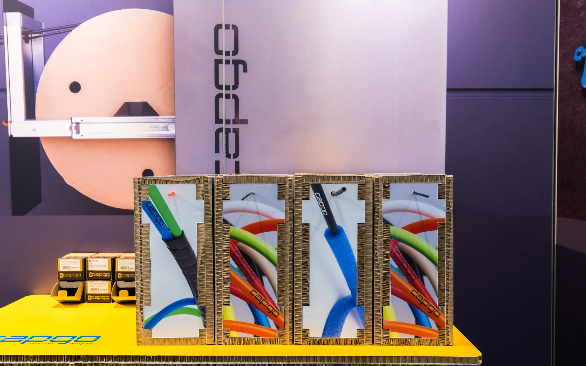 Vier Werkstattboxen mit bunten Außenhüllen stehen auf einem gelben Tresen am Messestand von capgo, dahinter eine violette Wand mit dem Schriftzug der Marke.