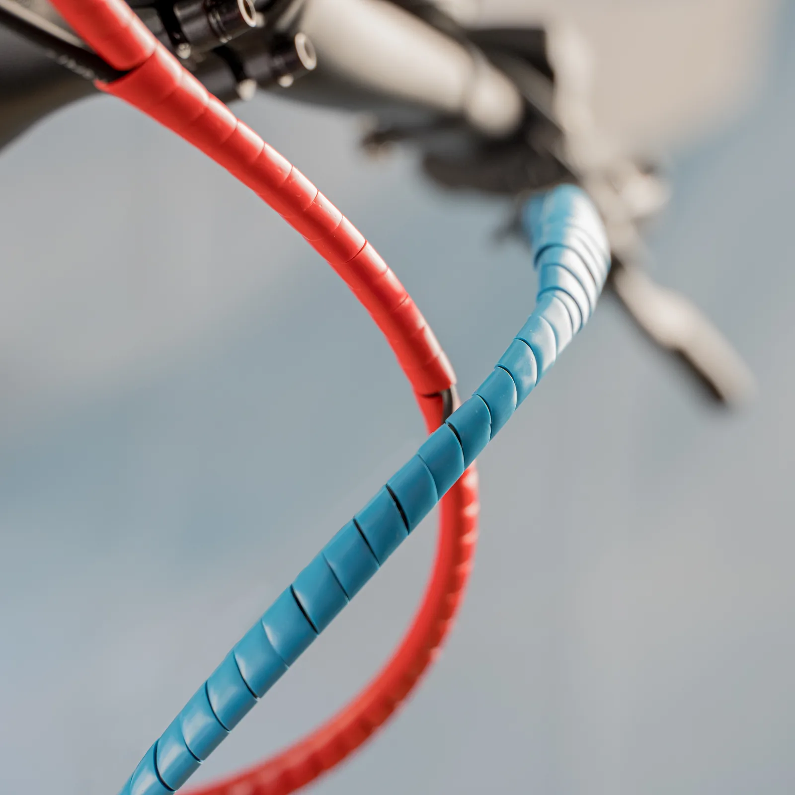 Ein roter und ein blauer Spiralschutz umwickeln die Züge an einem Mountainbike-Lenker vor hellblauem Hintergrund.