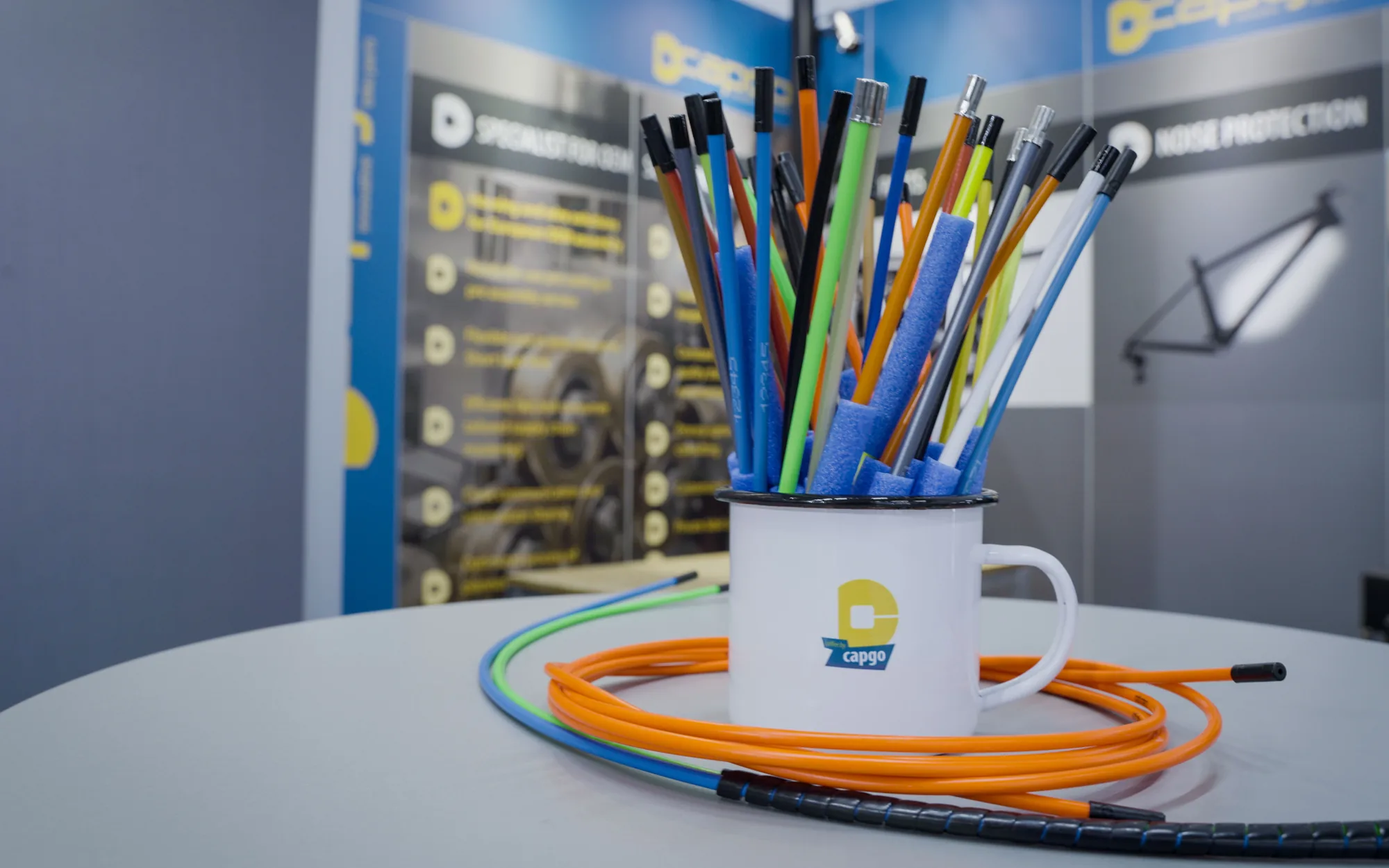 Ein weißer Emaillebecher mit dem capgo-Zeichen steht auf einem runden Tisch am Messestand, darin Dutzende Züge in Orange, Grün, Blau und Gelb, davor eine Schlaufe orangefarbener Außenhülle.