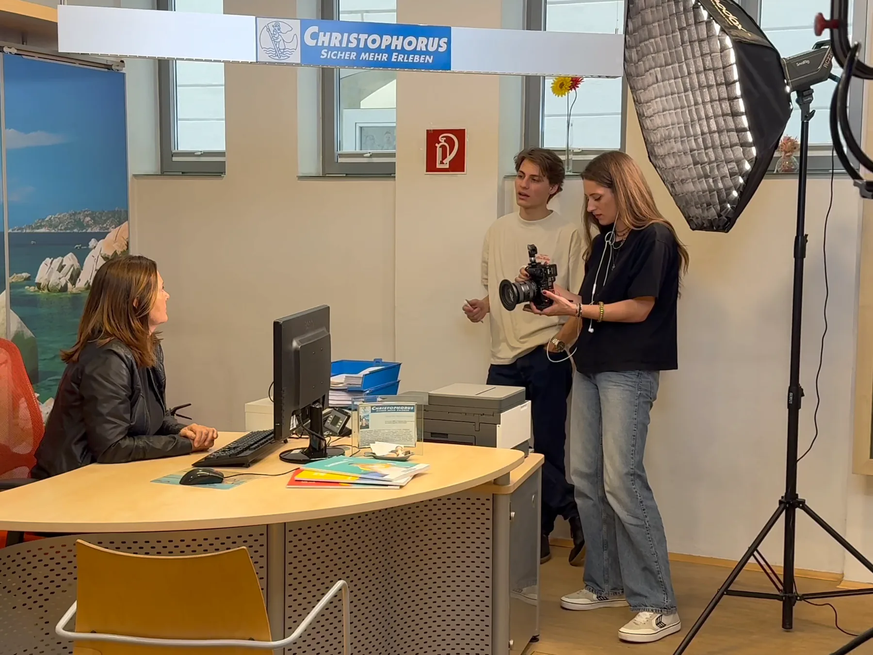 Beim Shooting im Reisebüro prüfen zwei aus dem Produktionsteam eine Aufnahme auf der Kamera, daneben eine Softbox auf dem Stativ, am Schreibtisch sitzt eine Beraterin, darüber das Schild Christophorus, Sicher mehr erleben.