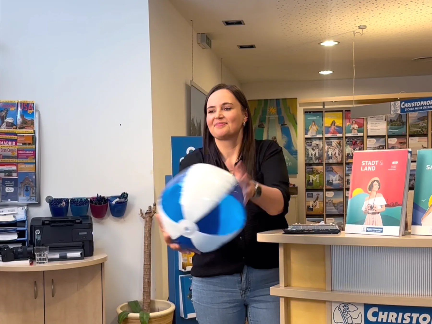 Eine Mitarbeiterin in schwarzer Bluse fängt beim Shooting an der Theke lachend einen blau-weißen Wasserball, hinter ihr die Prospektwand und der Katalog Stadt und Land.