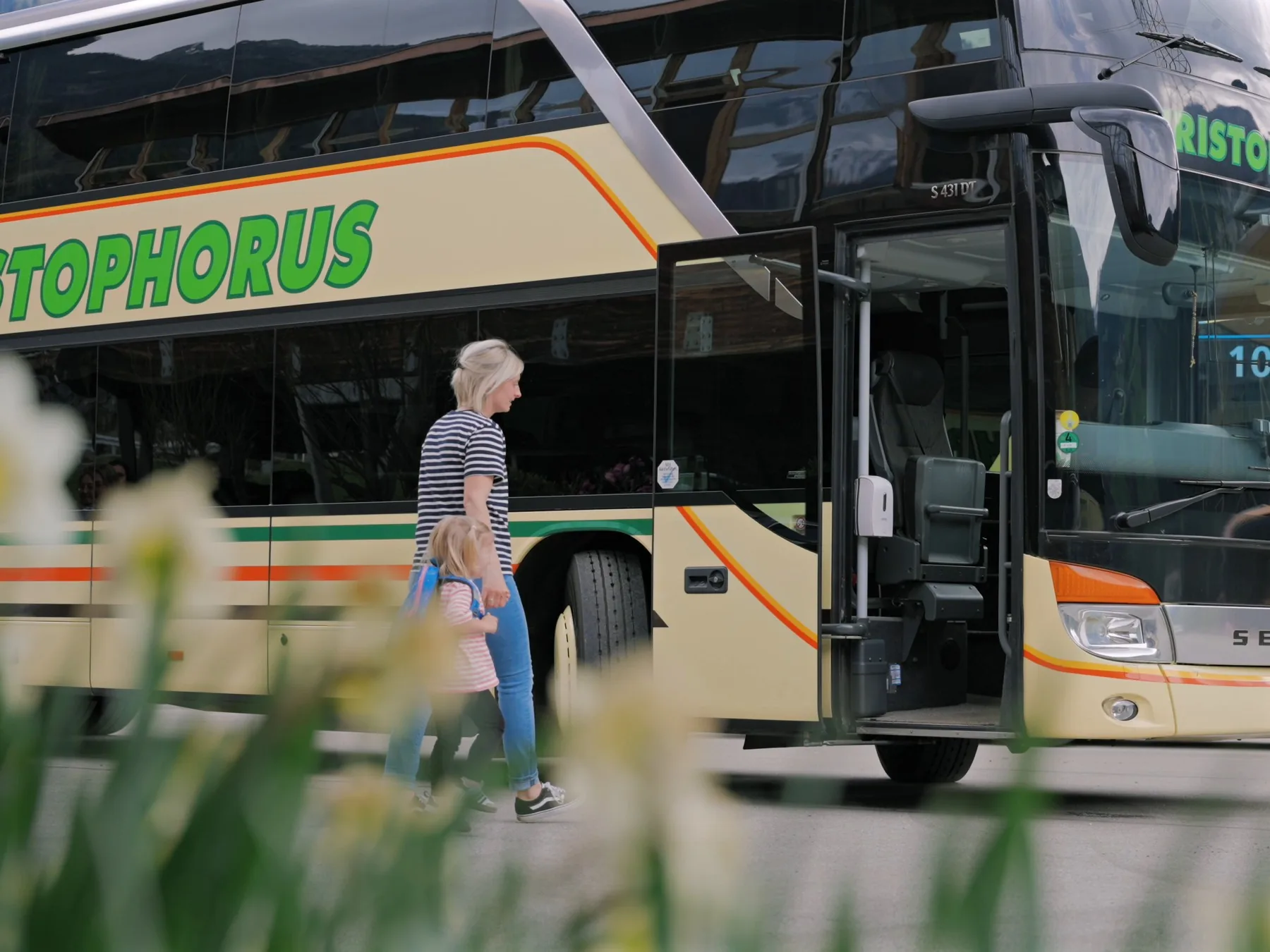 Eine Frau geht mit einem kleinen Mädchen an der offenen Tür eines Christophorus-Doppeldeckerbusses entlang, im Vordergrund unscharf weiße Narzissen.