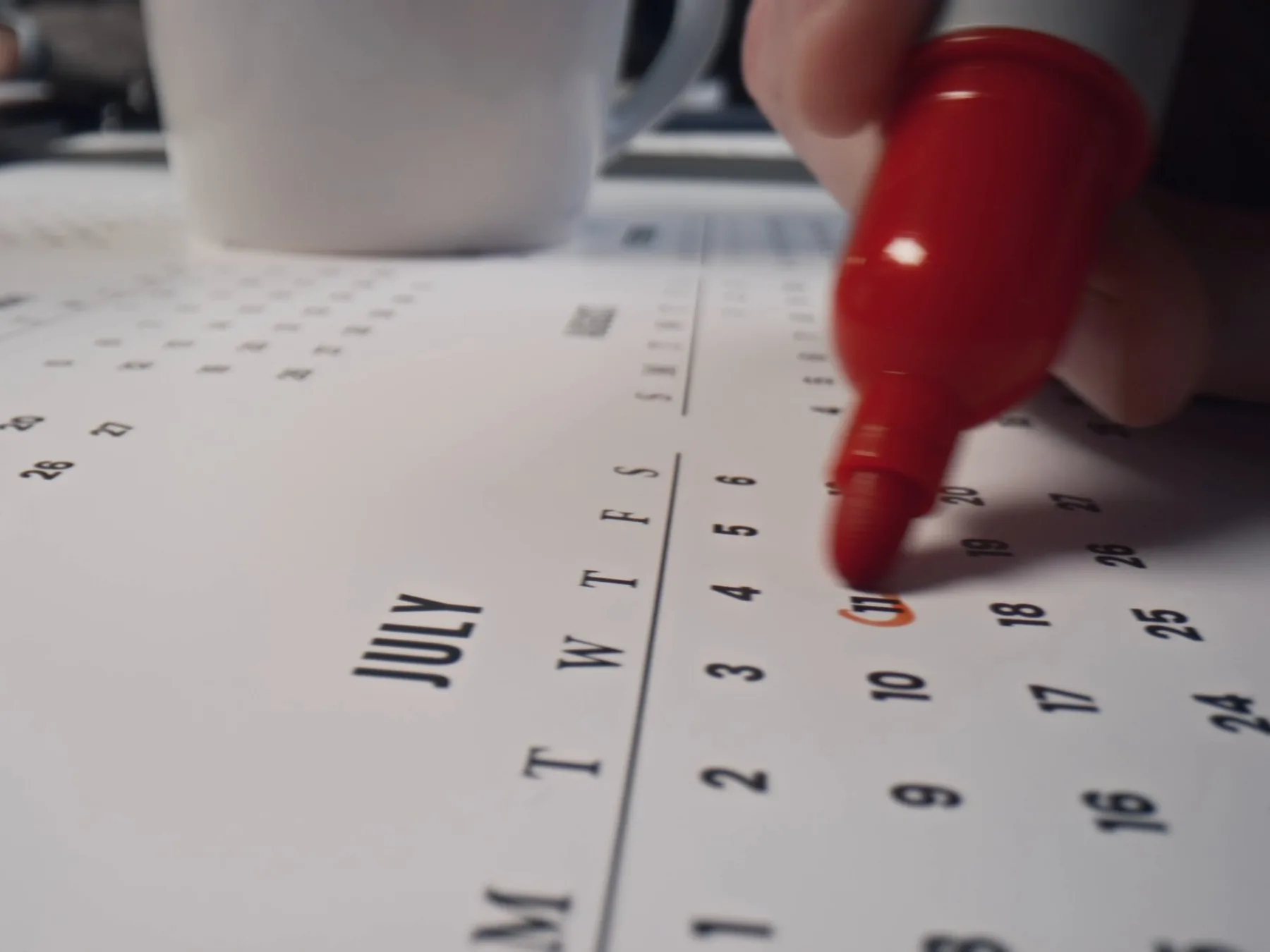 Eine Hand kreist mit einem roten Stift einen Tag im Juli auf einem Wandkalender ein.