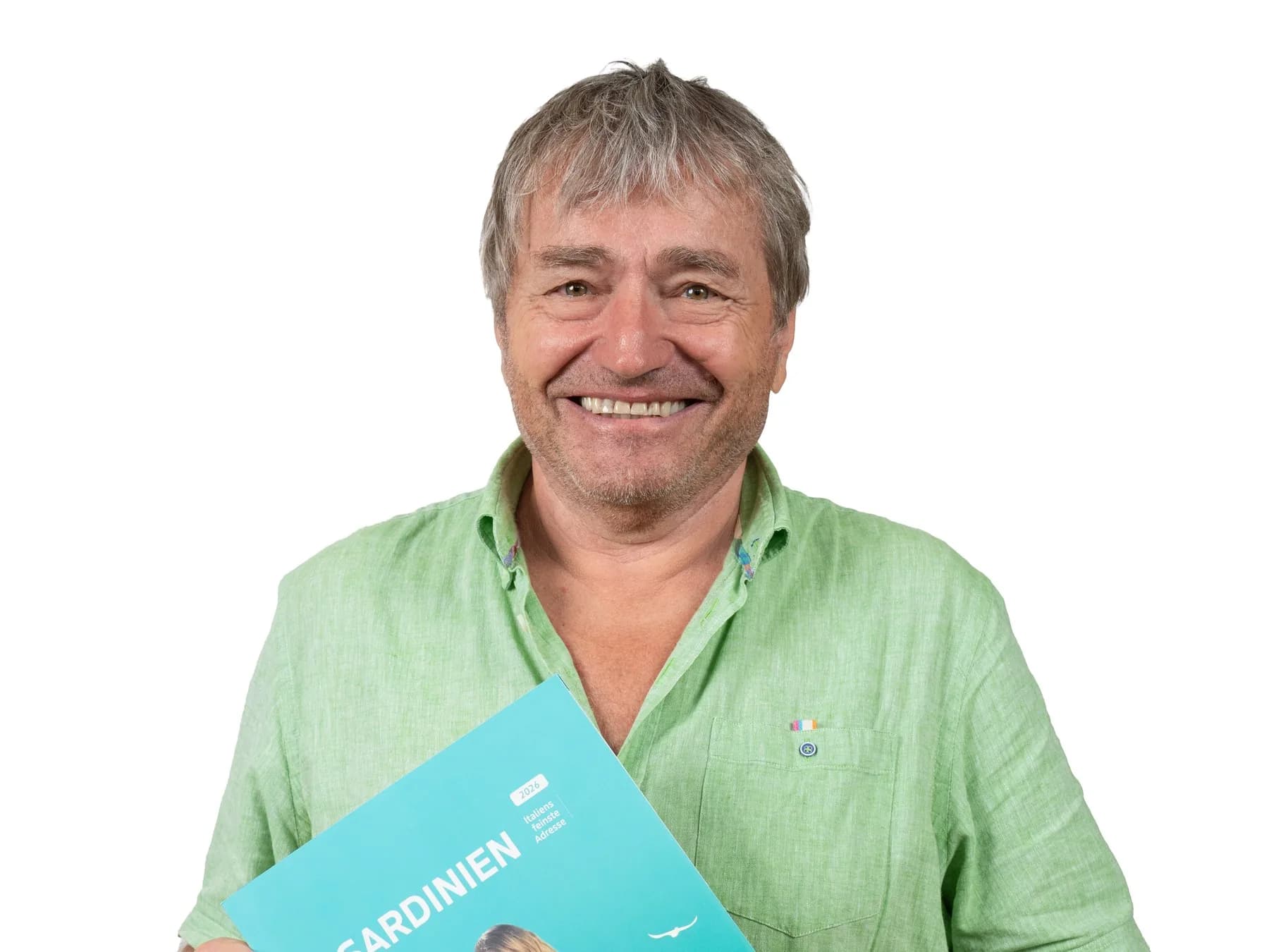 Andreas Kröll im grünen Leinenhemd lacht vor weißem Grund und hält den Christophorus-Katalog Sardinien vor die Brust.