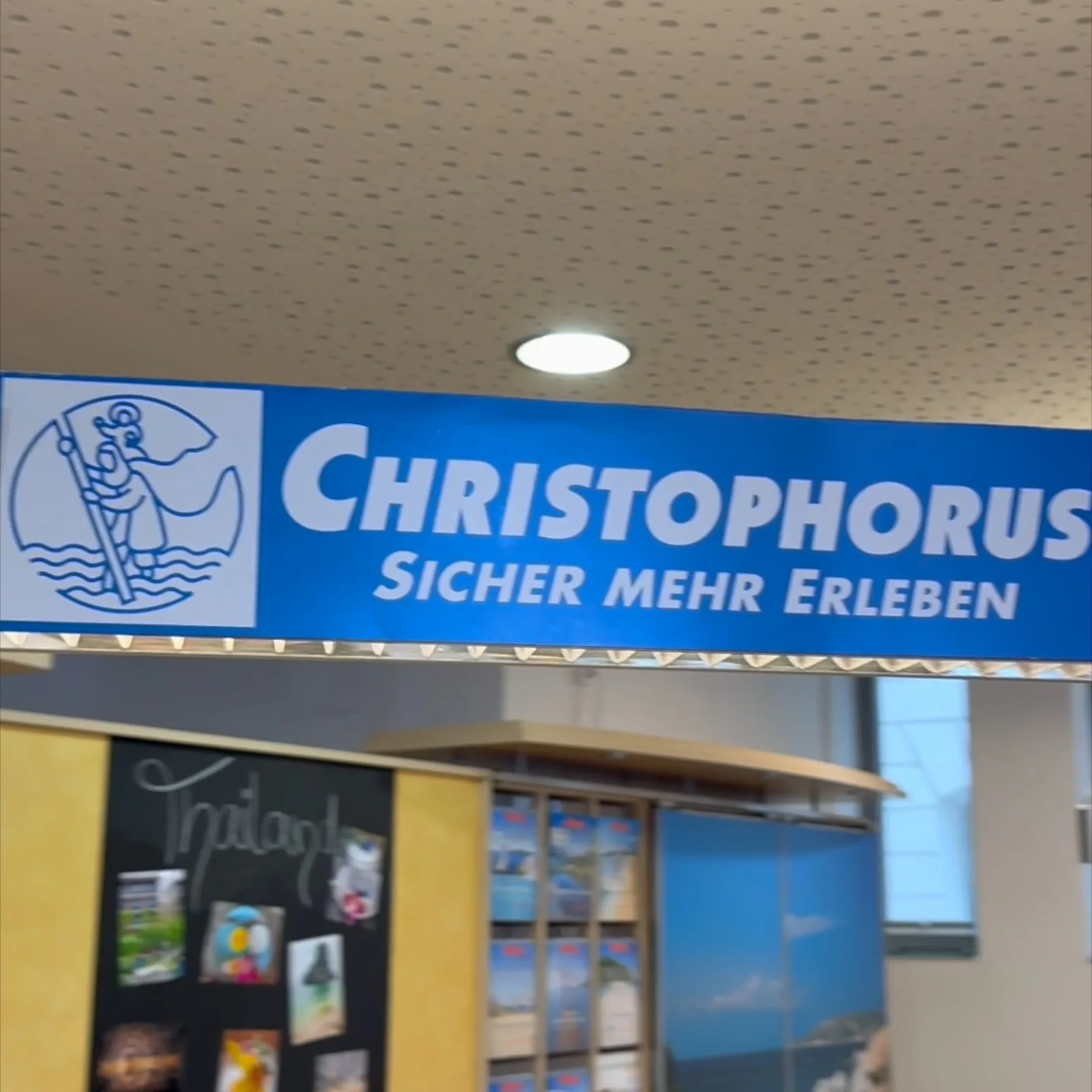 Das Schild Christophorus, Sicher mehr erleben, über dem Beratungsbereich eines Reisebüros, darunter eine Tafel mit Urlaubsfotos und die Prospektwand.